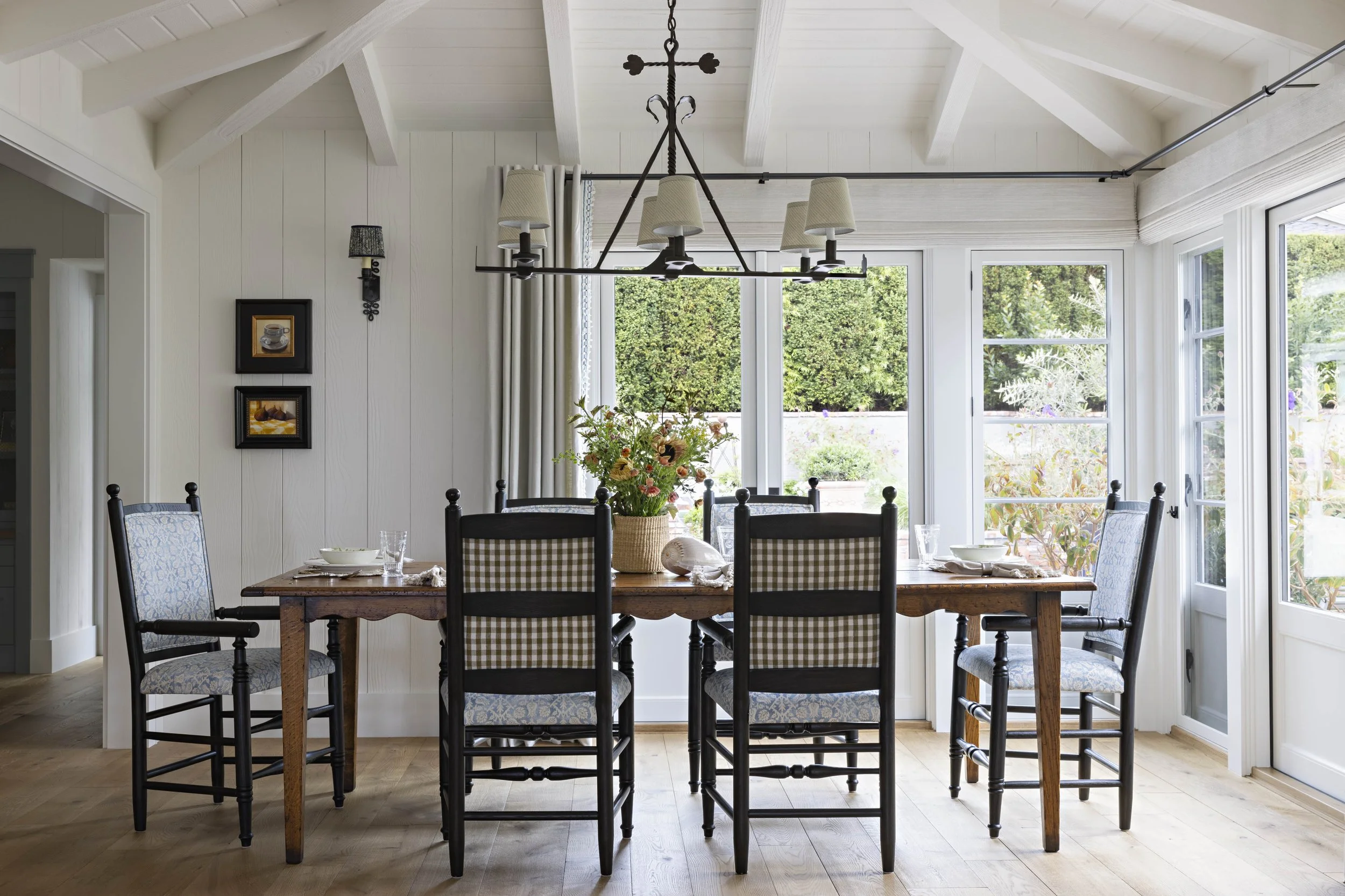 Dining room with a wooden table, six chairs with patterned upholstery, a flower arrangement, and large windows with green trees outside.