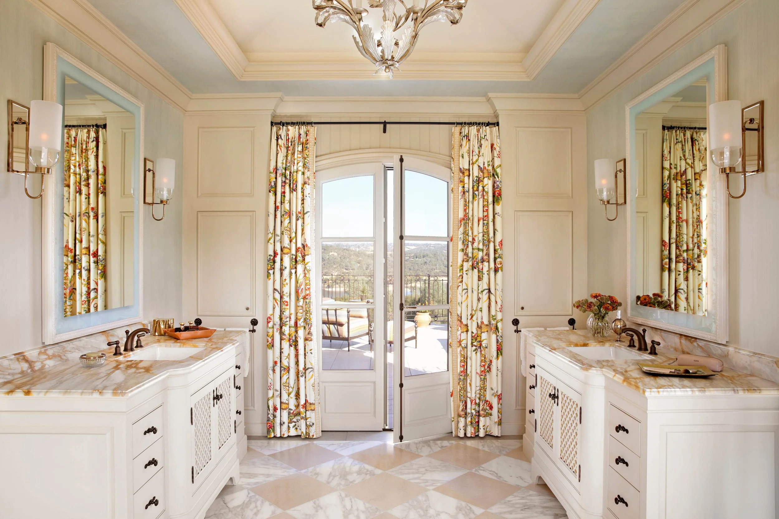 Bright bathroom with dual vanities, large mirrors, floral curtains, sliding glass doors leading to a balcony with outdoor furniture, chandelier, and light wall sconces.