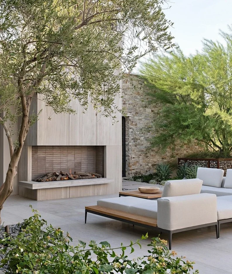 Modern outdoor patio with beige couches, a stone and wood fireplace, and lush green plants and trees.
