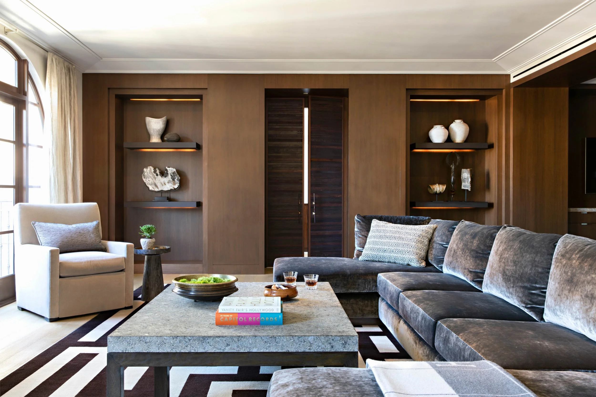 Modern living room with brown wall paneling, built-in shelves with decorative objects, a large gray velvet sofa, a light armchair, a stone coffee table with books and drinks, and large windows with curtains.