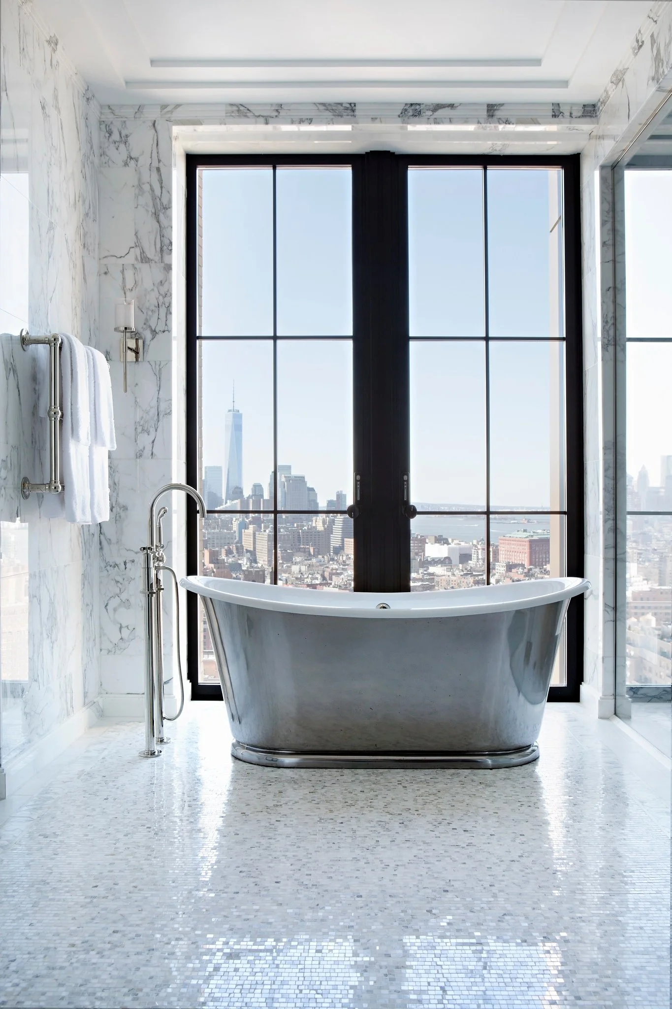 Luxurious bathroom with a freestanding bathtub in front of large floor-to-ceiling windows overlooking a city skyline.
