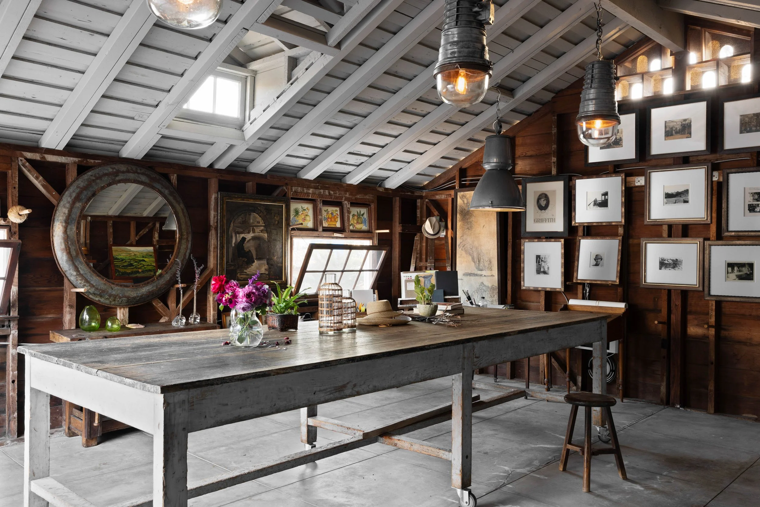 Interior of an art studio with a large wooden table in the center, decorated with flowers, hats, and decorative items; walls adorned with framed black-and-white photographs and artwork; industrial pendant lights hanging from the ceiling; rustic woode