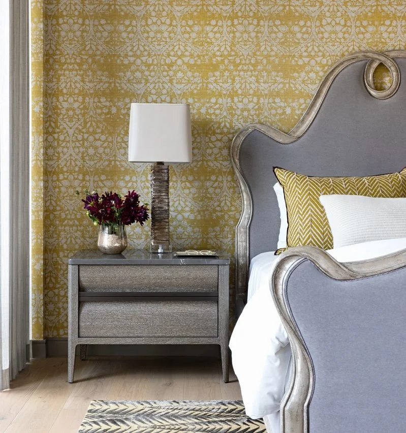 A bedroom corner with a gray upholstered headboard, a yellow patterned pillow, a bedside table with a silver vase holding purple flowers, a white lamp with a textured base, and a wall with yellow and white patterned wallpaper.