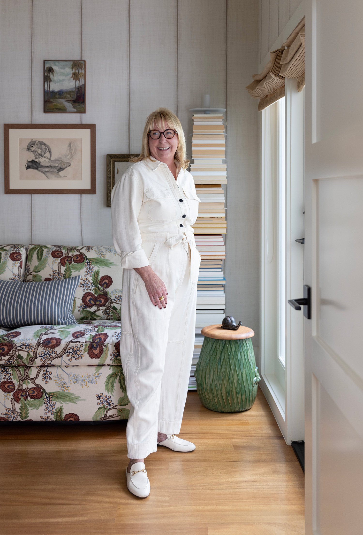 A woman with blonde hair, glasses, and a white outfit smiling inside a living room with a patterned sofa, a stack of books, and decor on the walls.