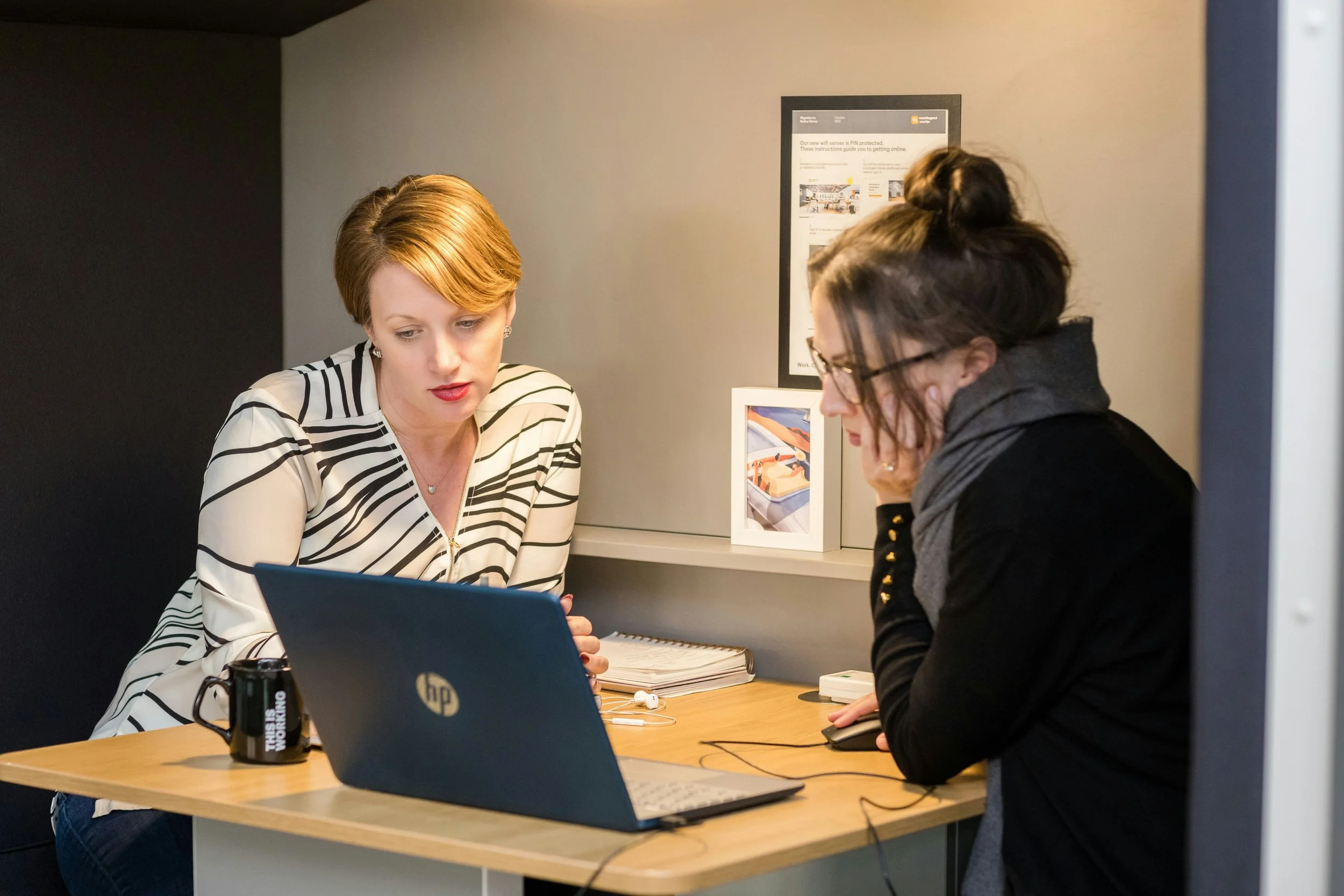 Two women sitting at a desk working on a laptop, one with glasses and dark hair in a bun, the other with short red hair, wearing a striped shirt.