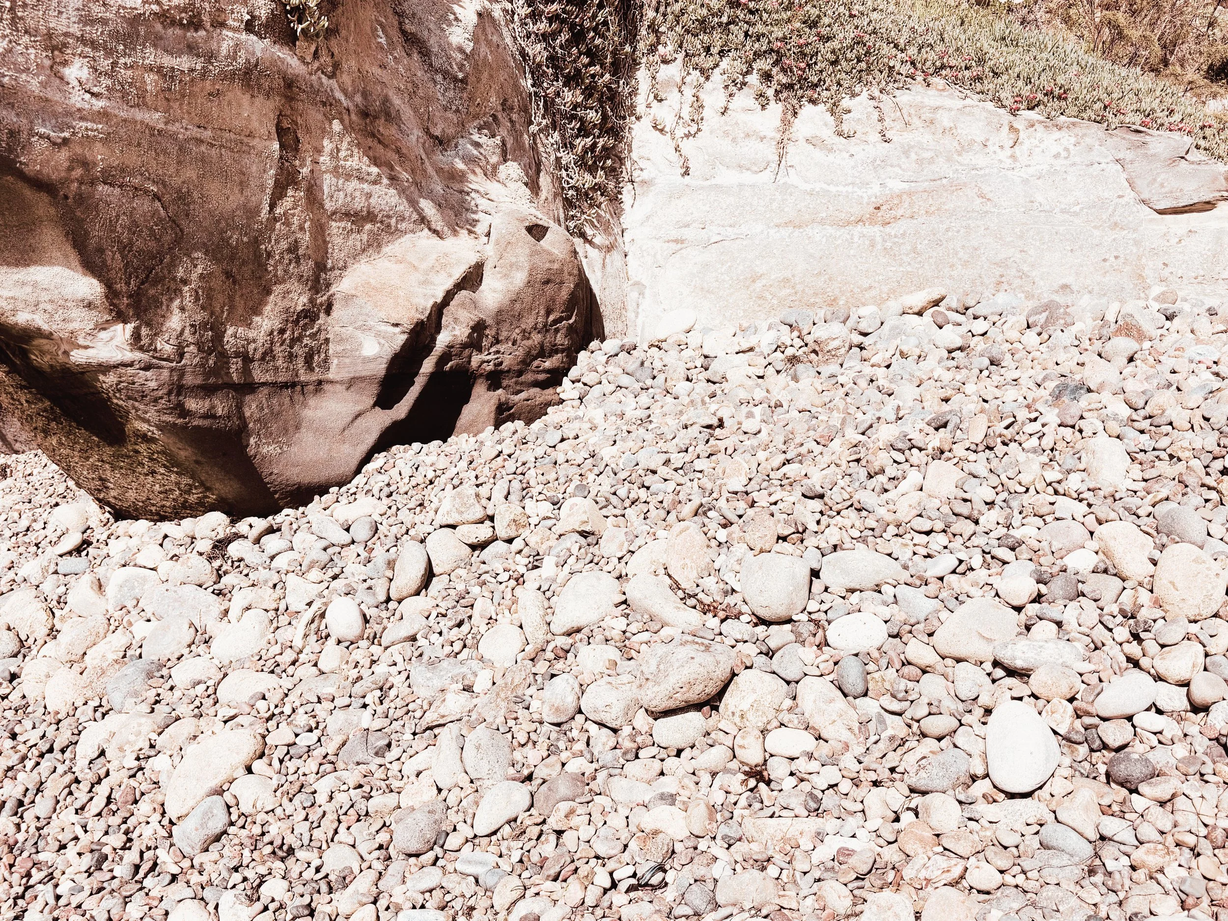 Close-up of a stone cliff with large boulders and small stones under bright sunlight, with a steep rock face and some sparse vegetation in the background.