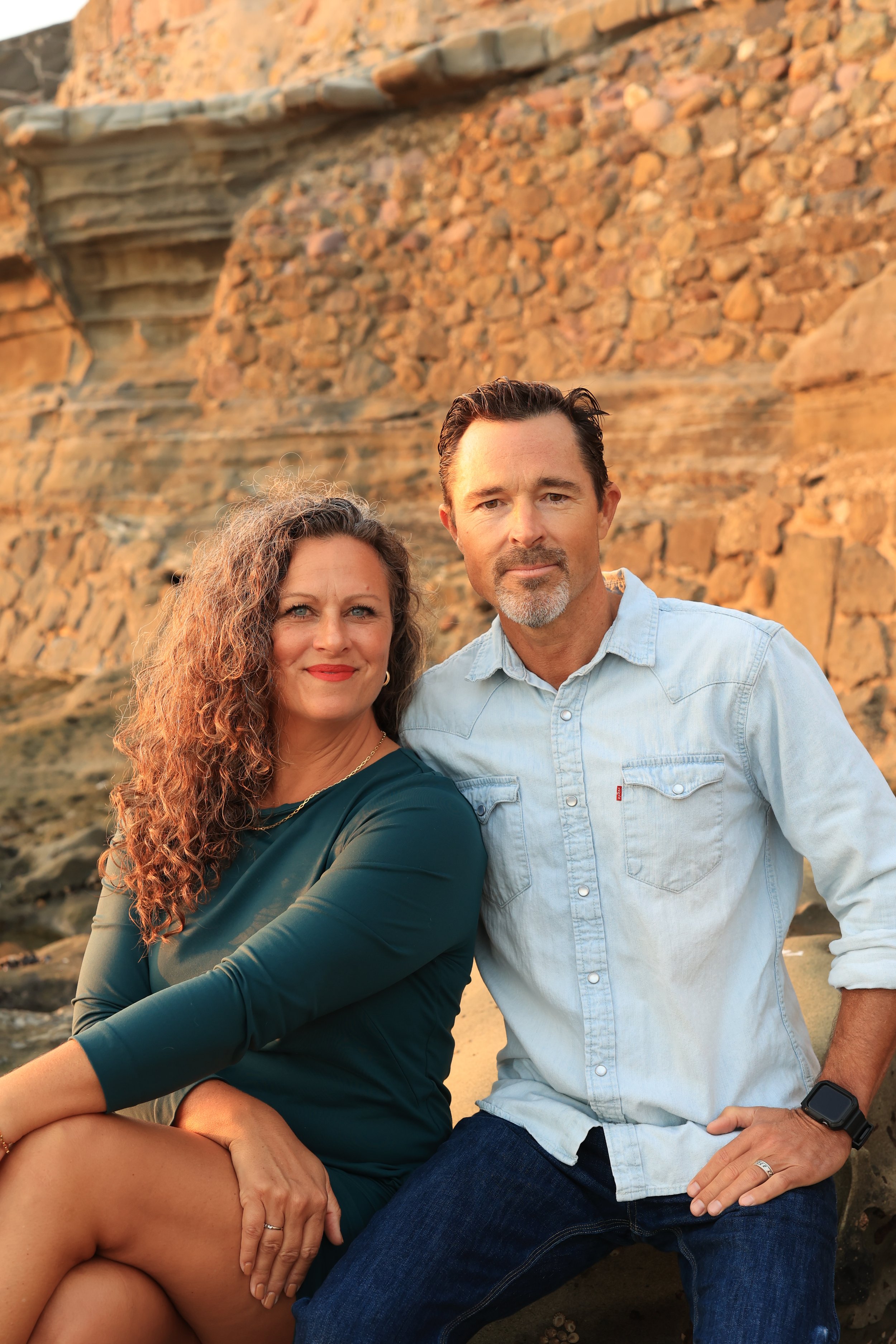 A man and woman sitting on rocks by a brick cliff at sunset.