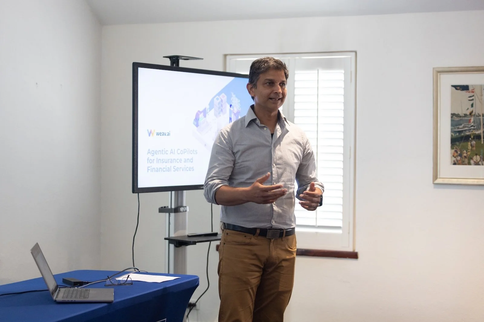 A man giving a presentation in a room with a large monitor displaying a slide that reads 'Agentic AI CoPilots for Insurance and Financial Services' with the logo 'weav.ai'.