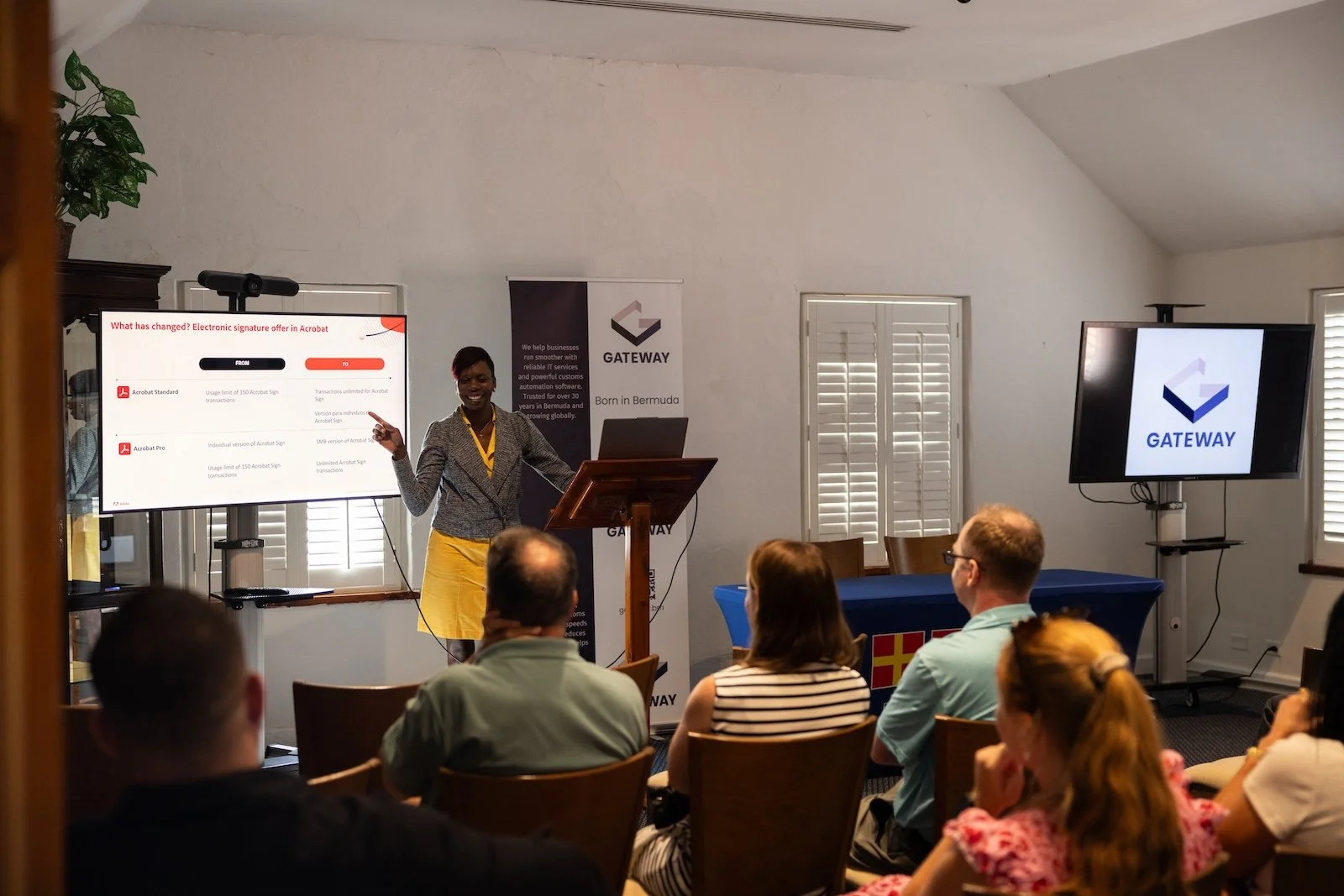 A woman presenting in front of a group during a business seminar. She is standing beside a large screen displaying a slide about electronic signature options in Acrobat. The room has white walls, the windows have white shutters, and there are two add