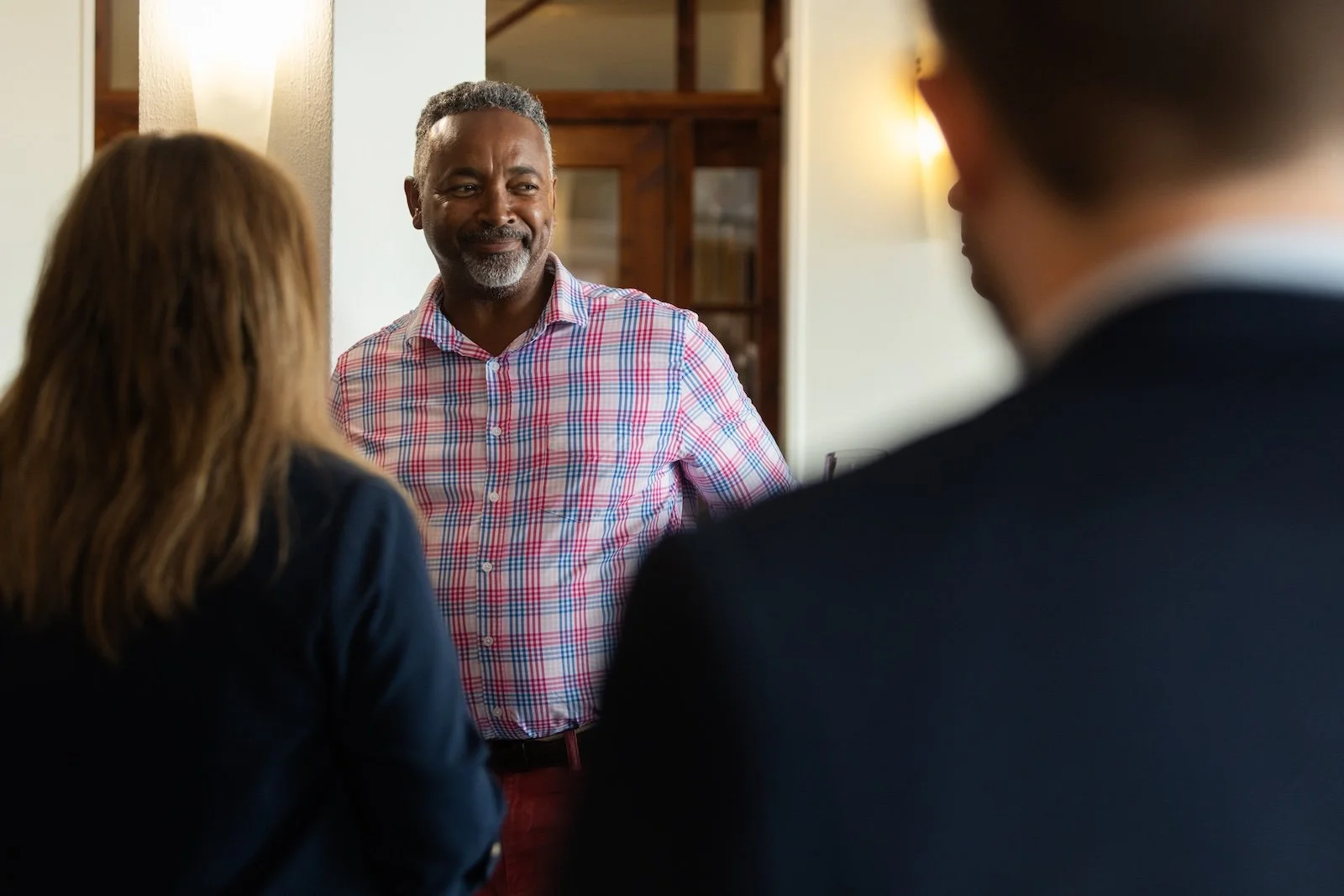 A man with gray hair and a beard, wearing a pink plaid shirt, smiling and talking to two people with their backs to the camera in an indoor setting with warm lighting.