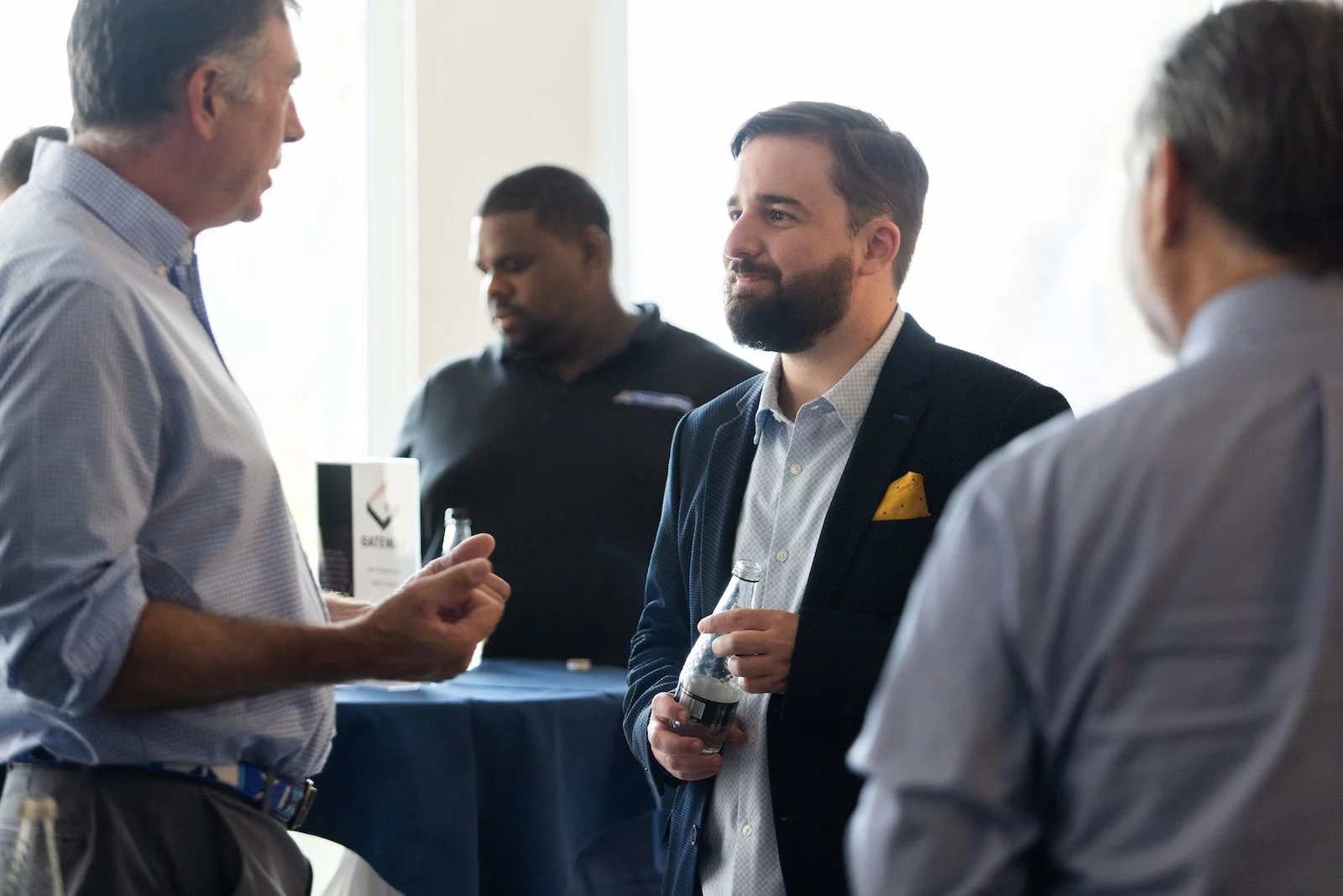 Group of four men in business casual clothes having a conversation at an indoor networking event, some holding beverages.