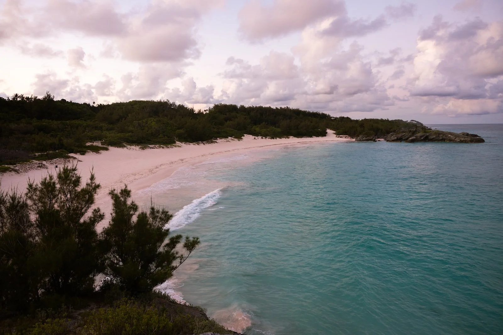 A scenic view of a sandy beach overlooking calm turquoise water, with lush green bushes and trees on the shoreline under a cloudy sky.