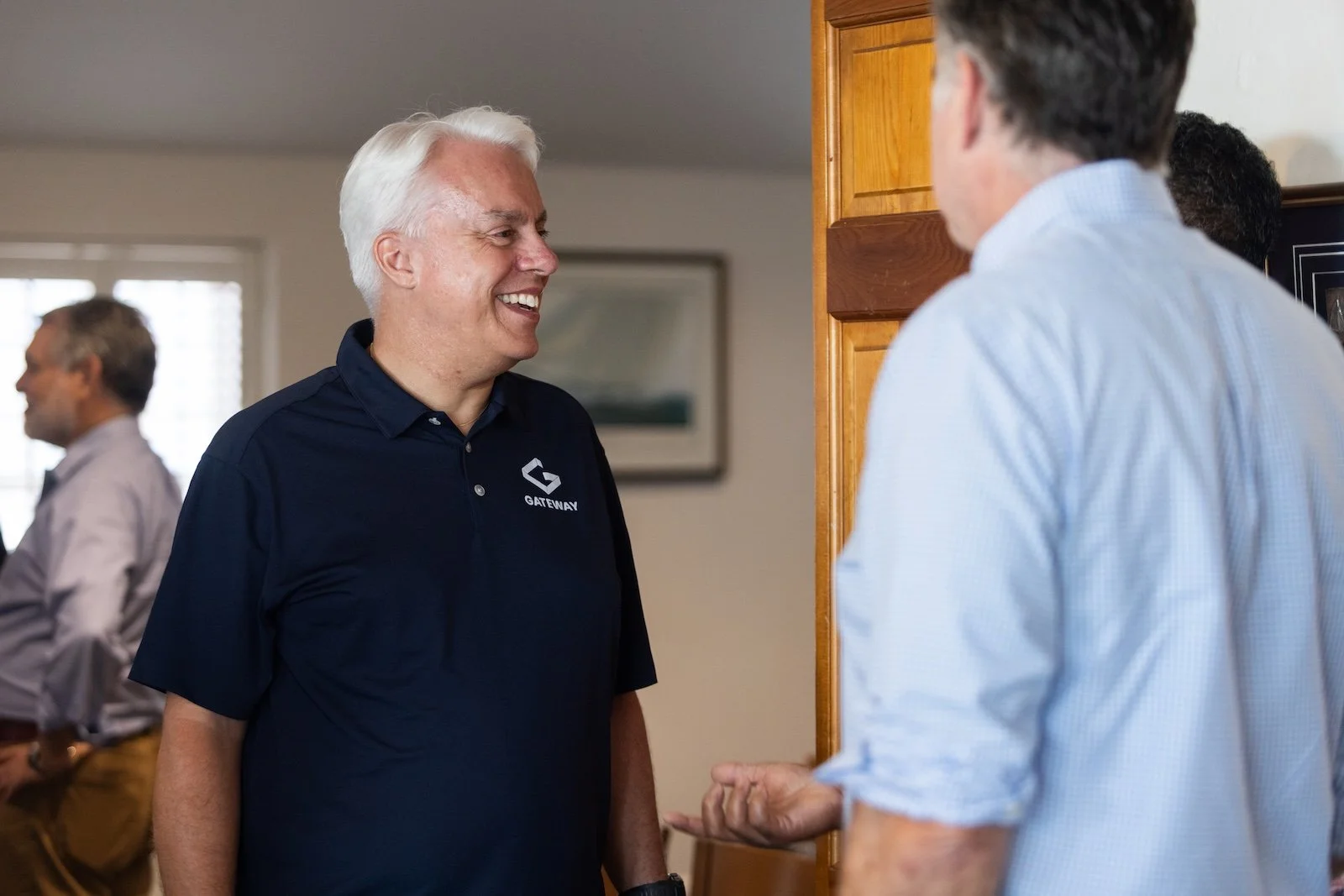 Two men smiling and talking to each other indoors, one wearing a navy blue shirt with a company logo, the other in a light blue shirt.