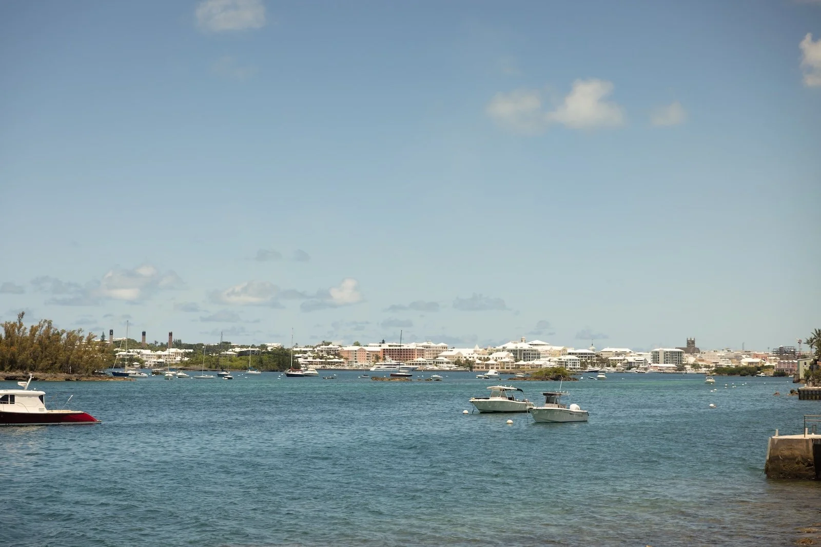 A calm waterway with anchored boats and a cityscape in the background under a blue sky with scattered clouds.