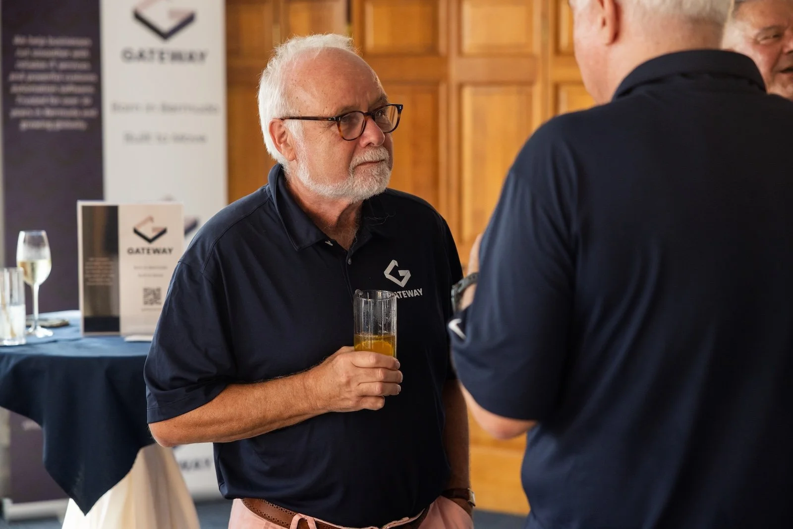 An older man with white hair, glasses, and a beard holding a glass of beer, talking to another man at a networking event with a Gateway banner in the background.