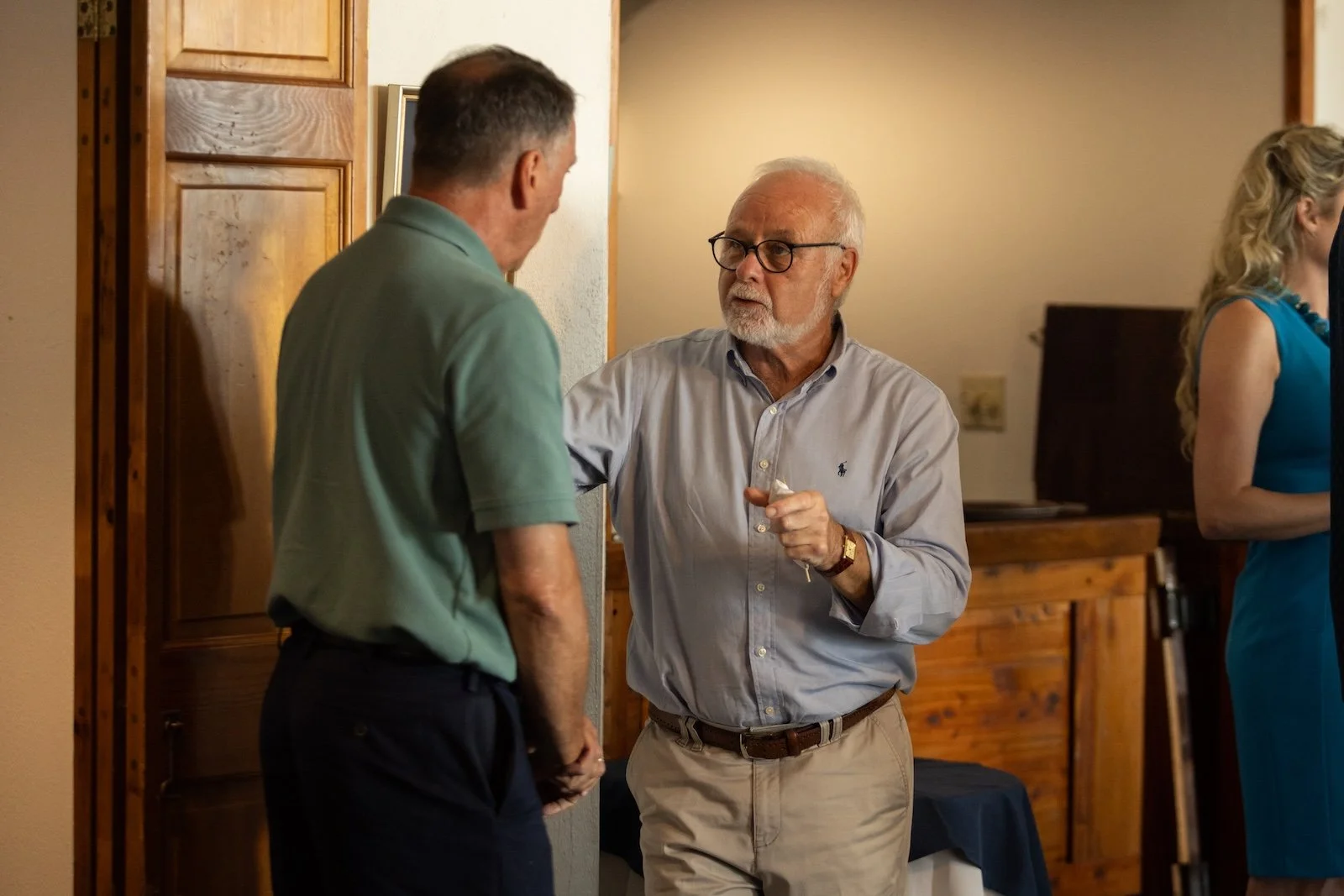 Two older men engaging in conversation at an indoor gathering, with a woman in a blue dress standing nearby.