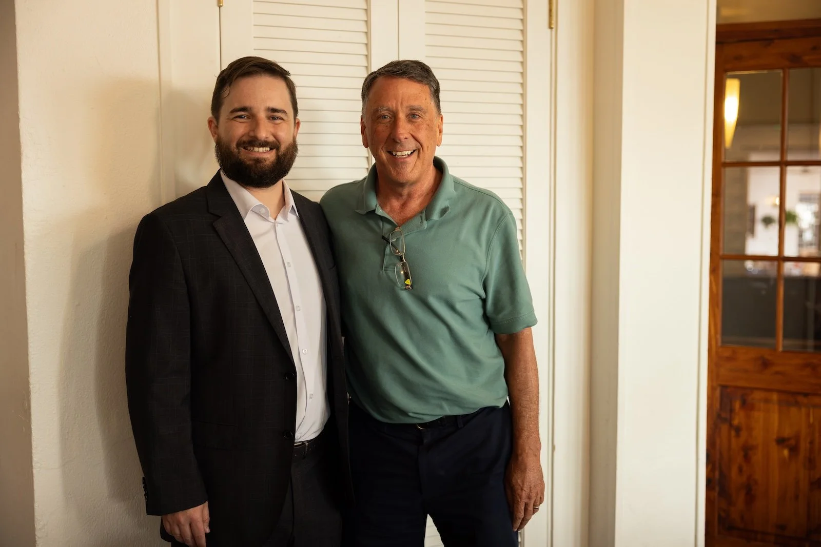 Two men smiling and standing together indoors, one in a dark suit and the other in a green polo shirt.