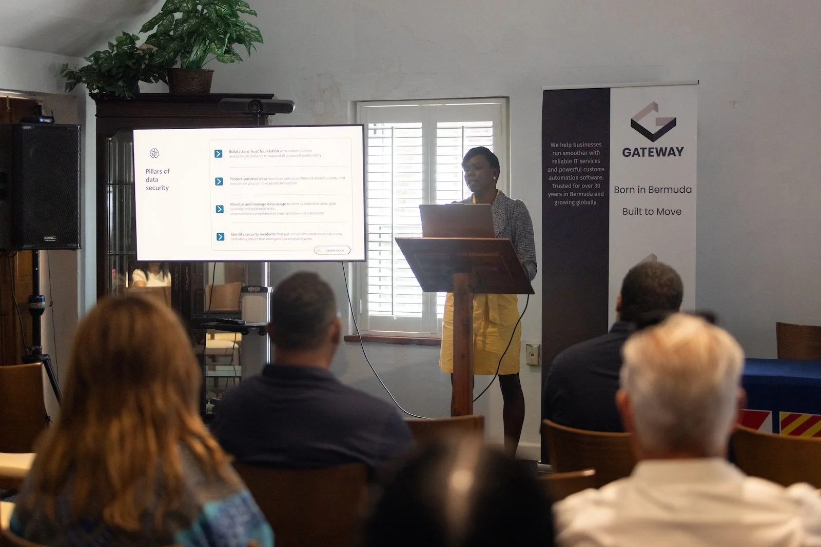 A woman giving a presentation in front of an audience, standing at a wooden podium with a laptop, with a large screen displaying presentation slides on data security. There are several people seated and watching the presentation inside a room with a