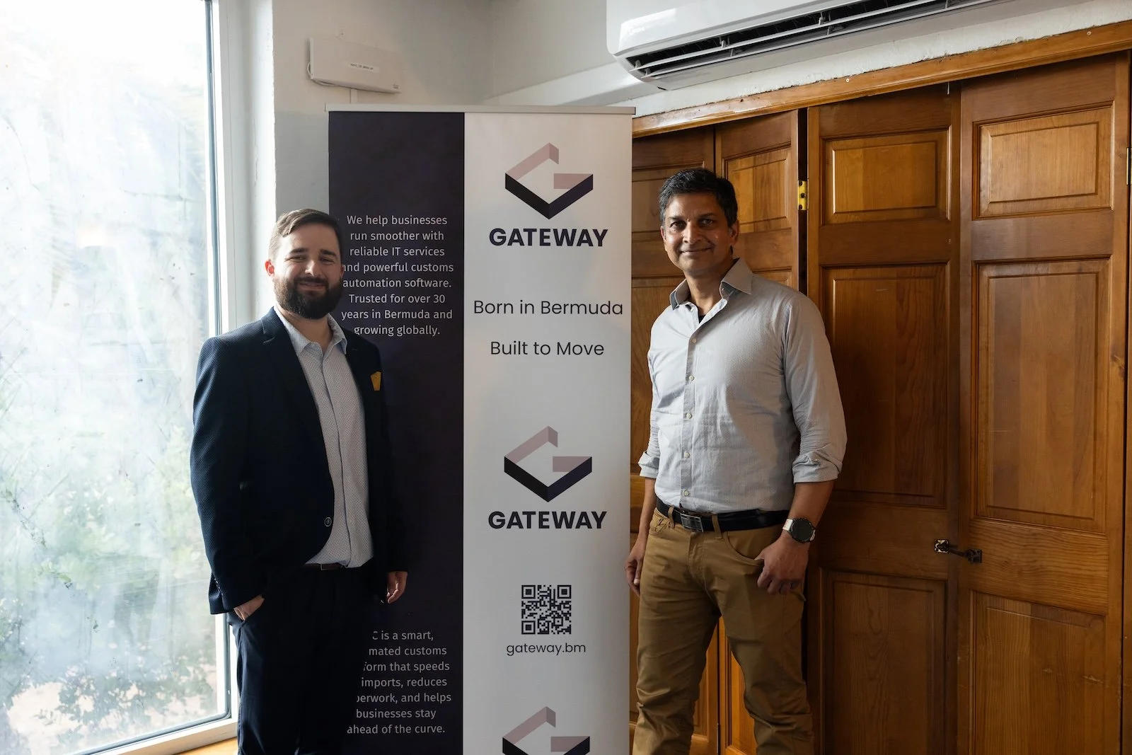 Two men standing indoors next to a vertical banner promoting Gateway, a company founded in Bermuda that offers IT services and customs automation software. The banner includes the Gateway logo, slogan 'Born in Bermuda, Built to Move,' a QR code, and