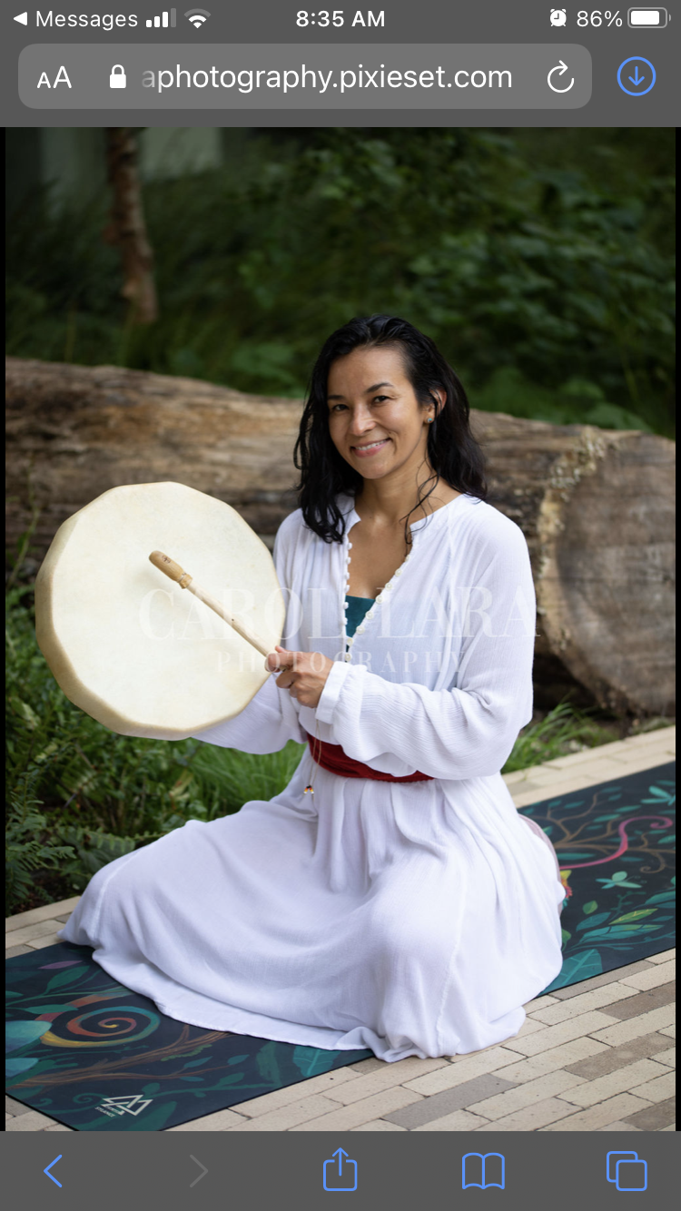 A woman with dark hair wearing a white dress and red belt, sitting on a colorful yoga mat outdoors, holding a large drum and a drumstick, surrounded by green foliage and trees.