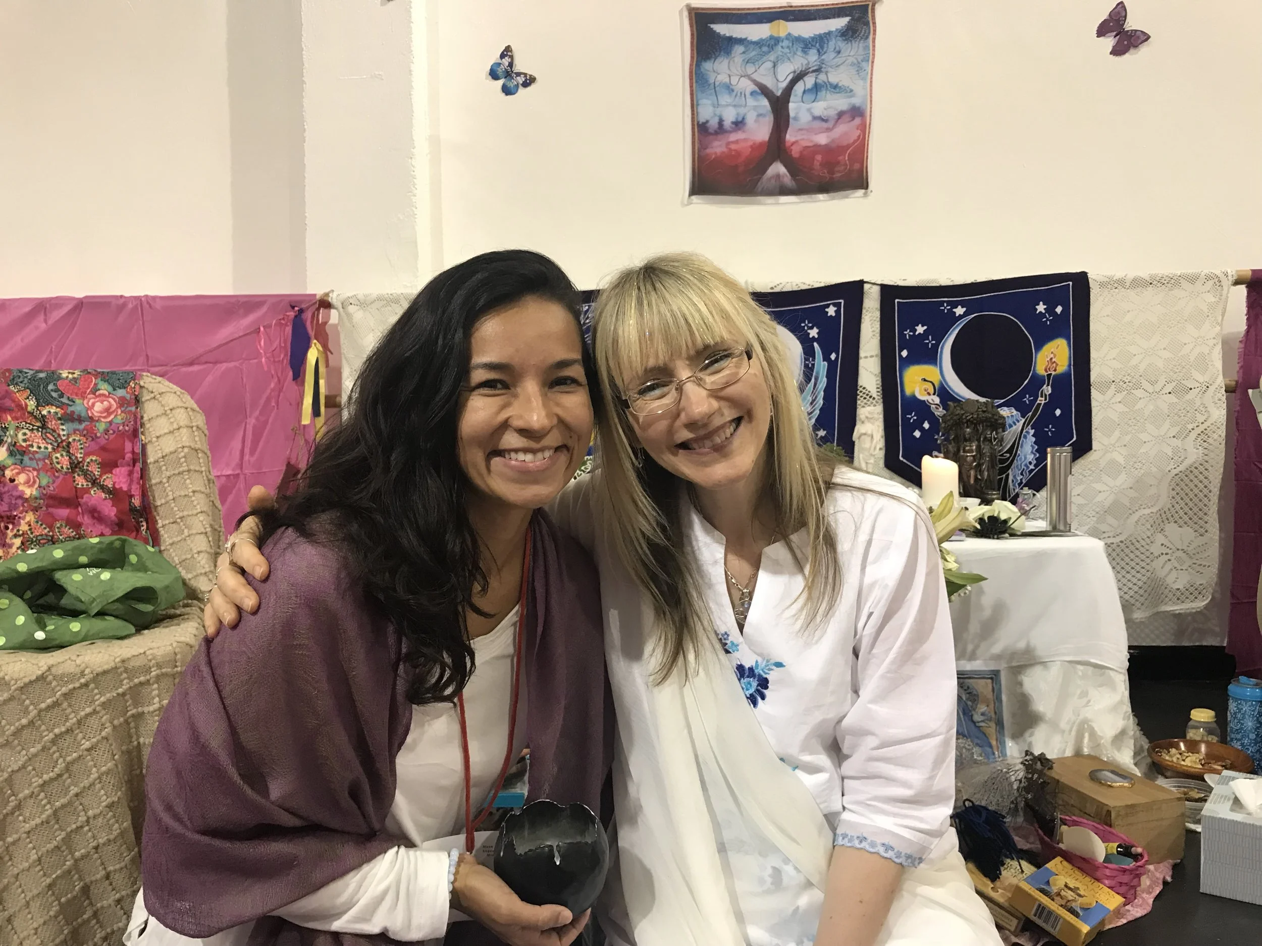 Two women smiling and sitting close together in a decorated room, with spiritual and artistic decorations in the background, including a moon and tree tapestry, candles, and flowers.