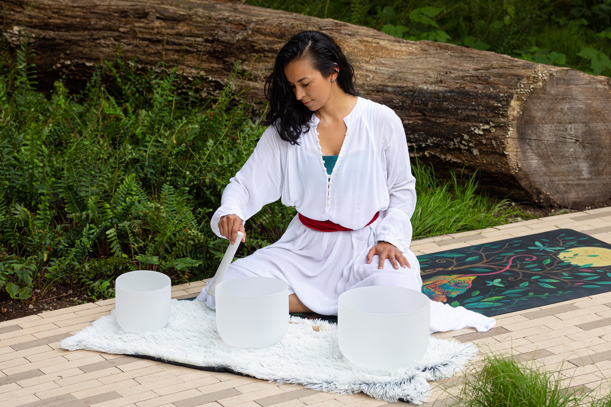 Woman in white dress sitting outdoors on a blanket, playing crystal singing bowls.