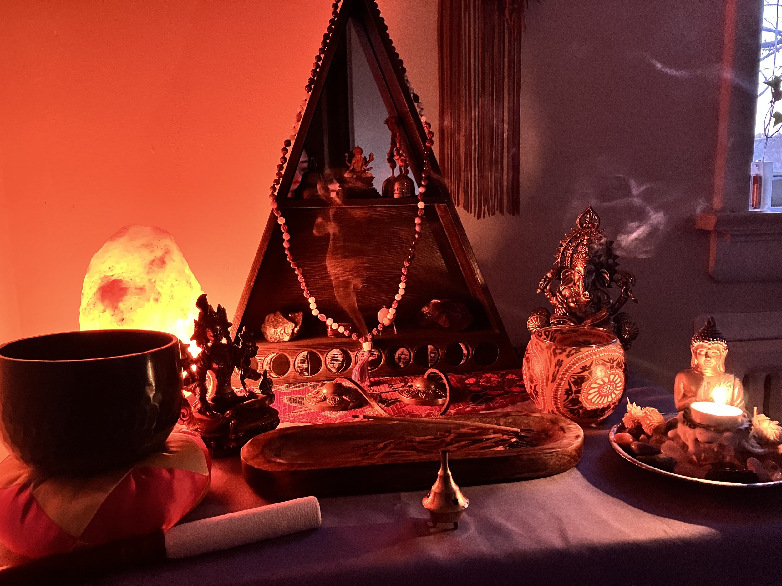 A spiritual altar with various statues, including Lord Ganesha and Buddha, a Himalayan salt lamp, a Himalayan salt candle holder, incense, and decorative objects, illuminated by soft red and warm lighting.