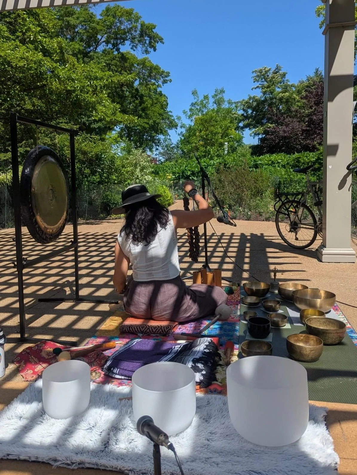 A woman sits outdoors on a blanket with singing bowls and various percussion instruments, holding a mallet. She wears a wide-brimmed hat and a white sleeveless top. A gong is visible to her left, and a bicycle is parked nearby. The scene is shaded with sunlight creating a pattern of shadows on the ground, with trees and blue sky in the background.