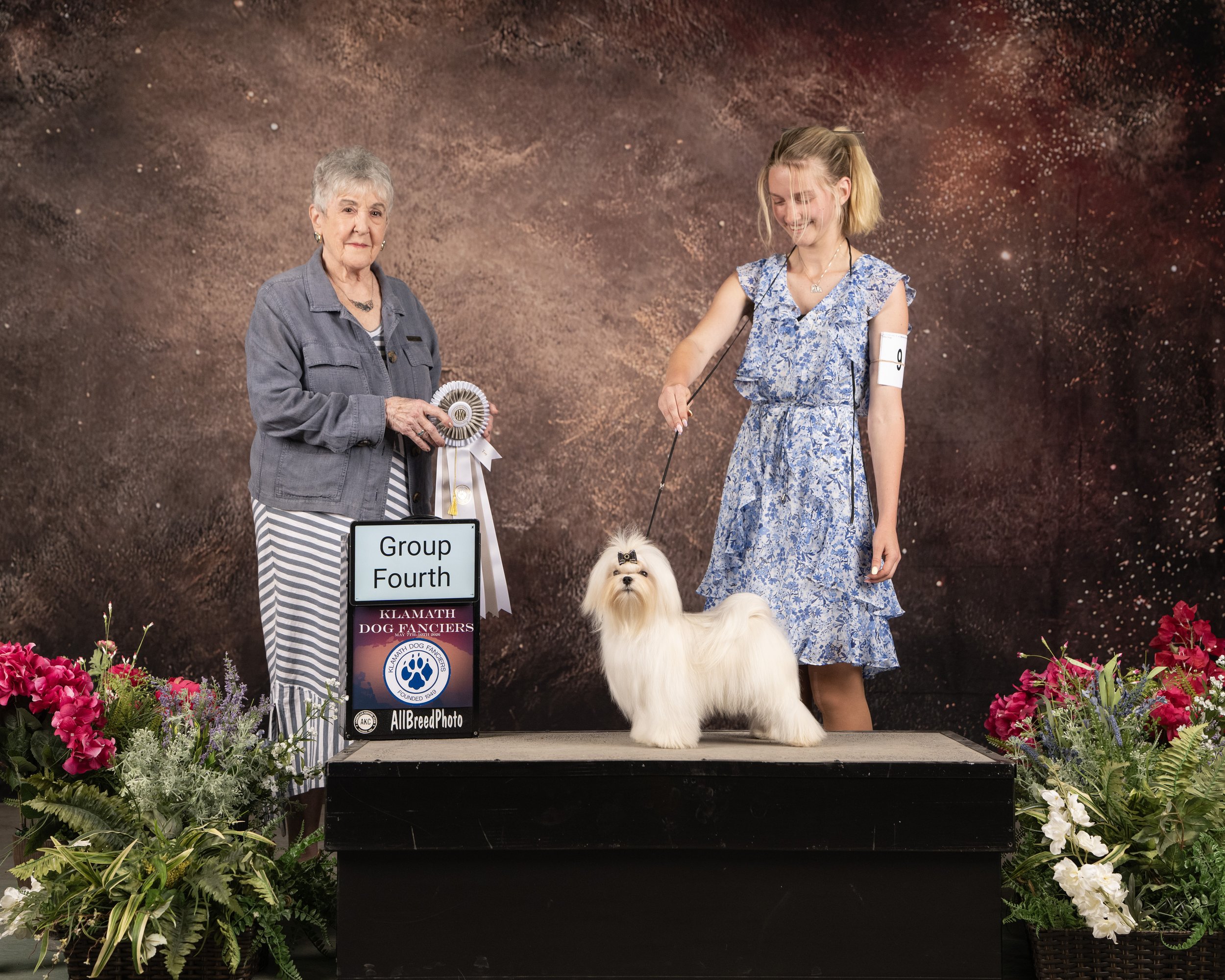 Two women with dog at a dog show, one holding a rosette ribbon, flowers around the stage, and a sign that reads 'Group Fourth' and 'Klamath Dog Fanciers.' The dog, a fluffy light-colored breed, stands on the stage.