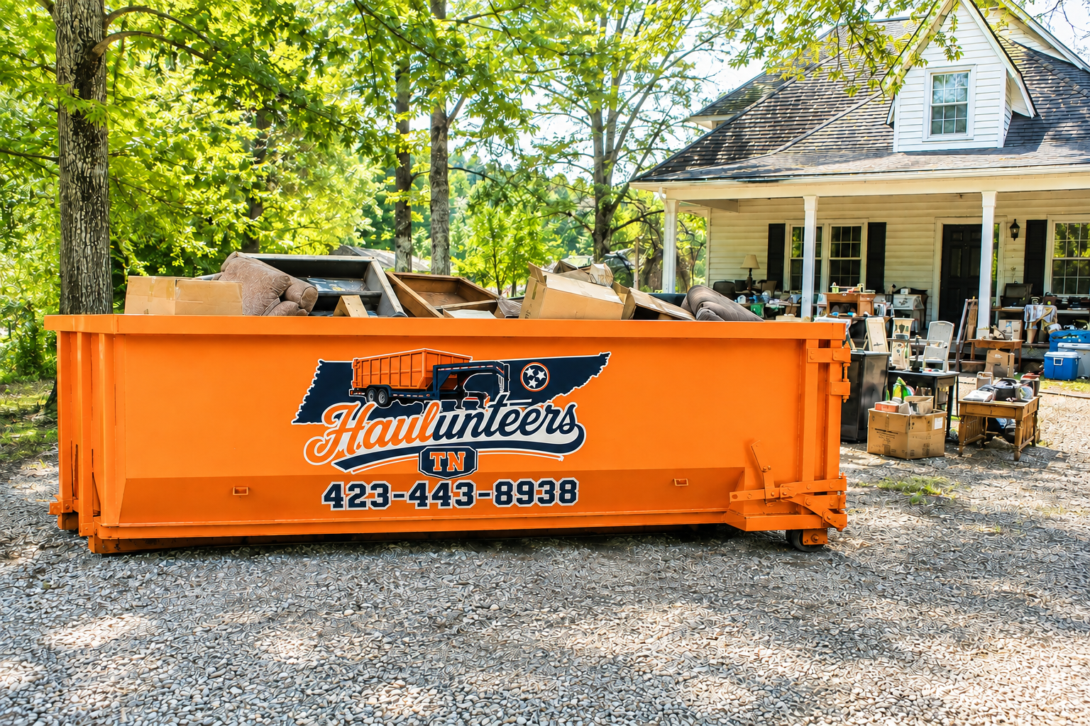 Orange dumpster filled with furniture and boxes in front of a house with trees and an outdoor seating area.
