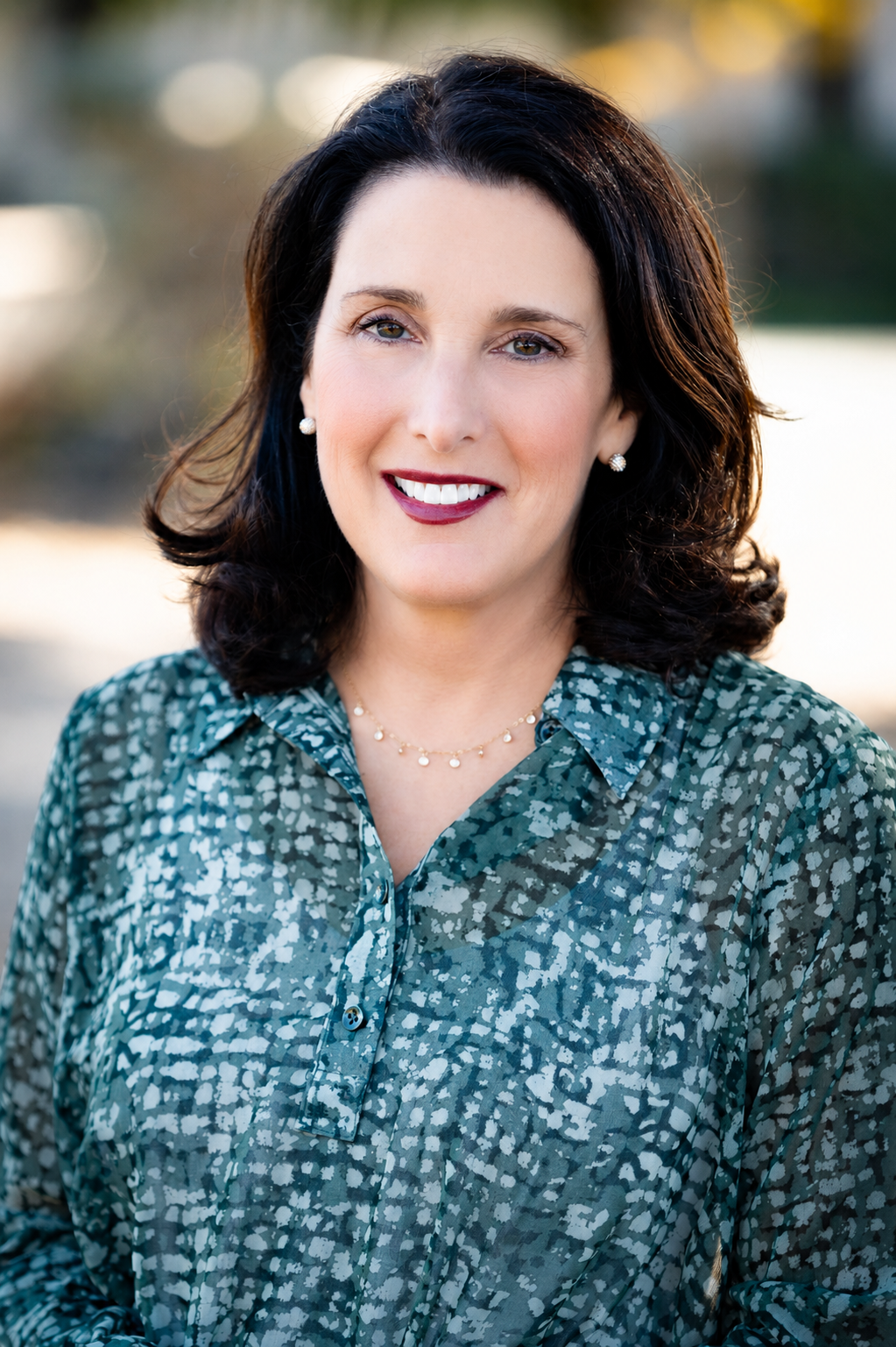 A woman with shoulder-length dark hair and light skin smiling outdoors, wearing a patterned blouse and pearl jewelry.