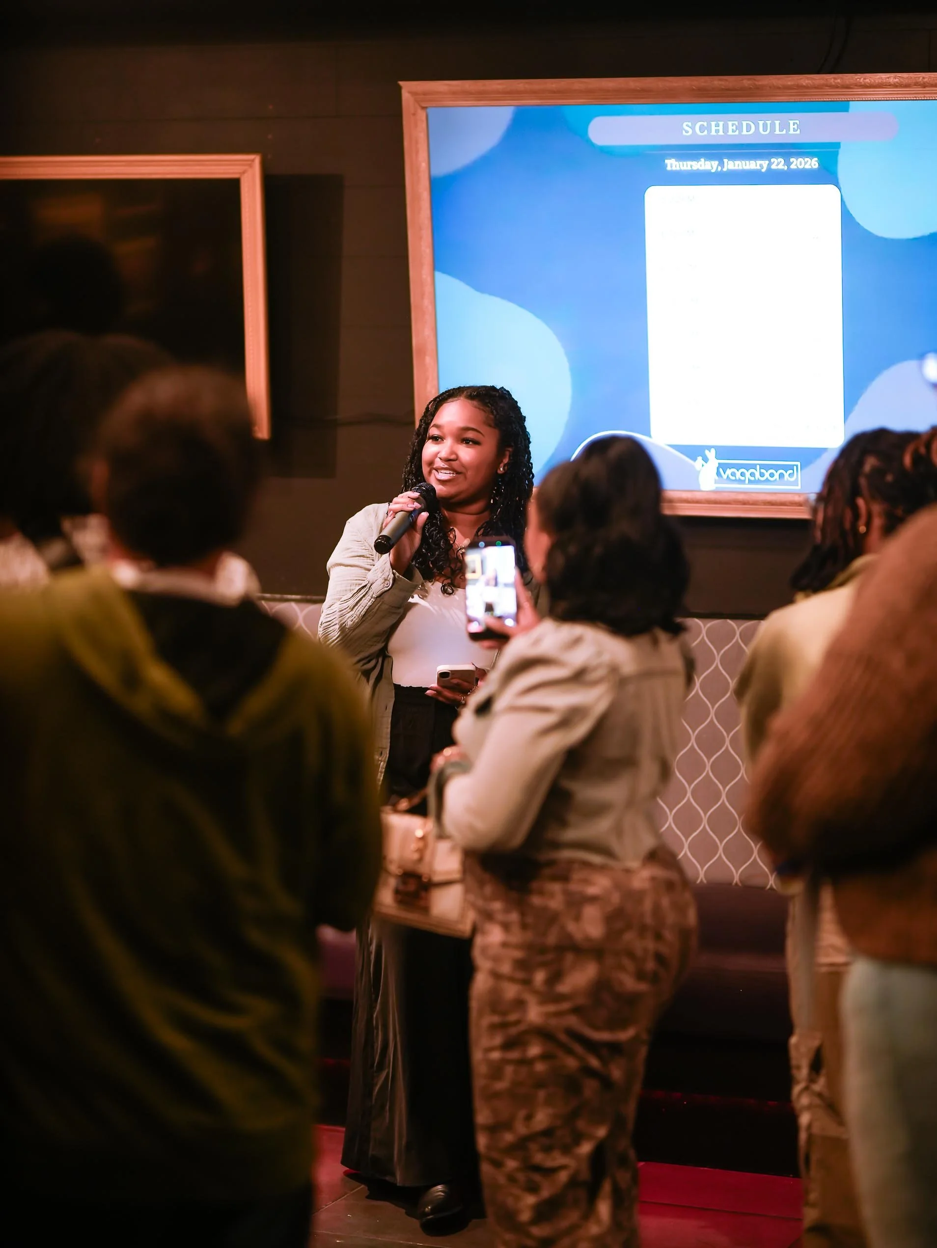 A woman is speaking into a microphone in front of an audience, with a large screen displaying a schedule behind her. The setting appears to be a presentation or being part of an event.