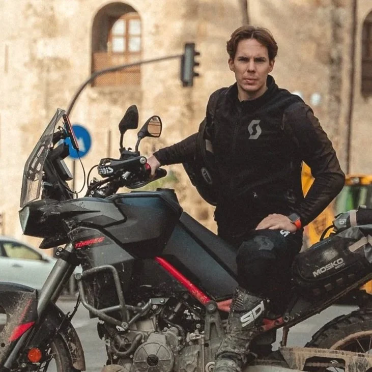 Uomo in abbigliamento da moto seduto su una motocicletta nera, in strada con sfondo di edifici antichi e semafori.