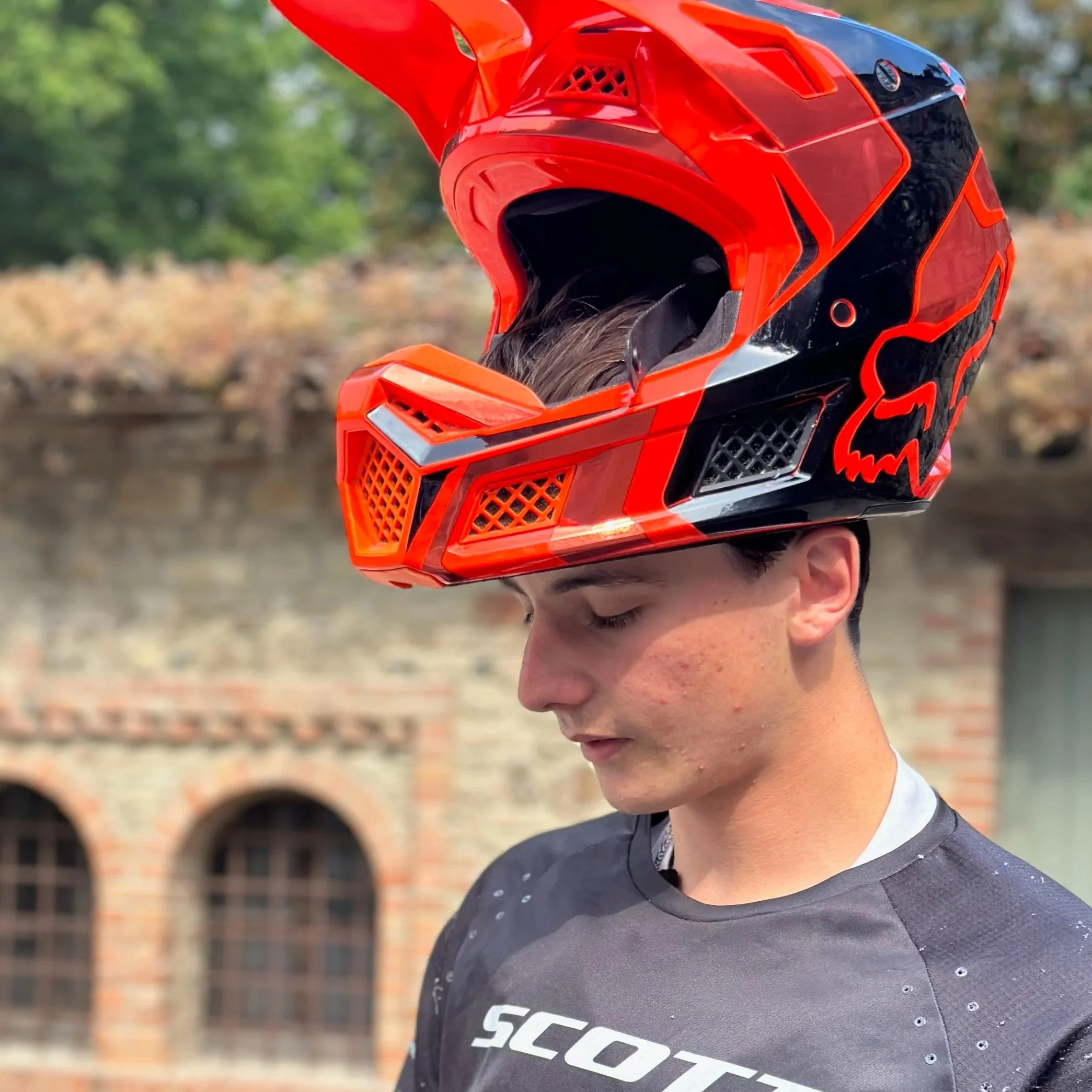 Giovane con casco da motocross rosso e nero, indossa una maglietta grigia, con sfondo di un edificio in mattoni e alberi verdi.