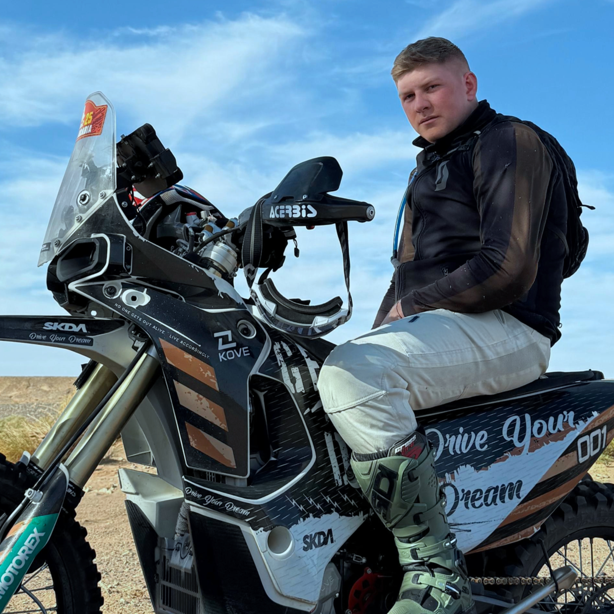 Giovane uomo seduto su una motocicletta da fuoristrada in un paesaggio desertico, indossando un giubbotto nero, pantaloni chiari e stivali da motocross, sotto un cielo azzurro con nuvole sparse.