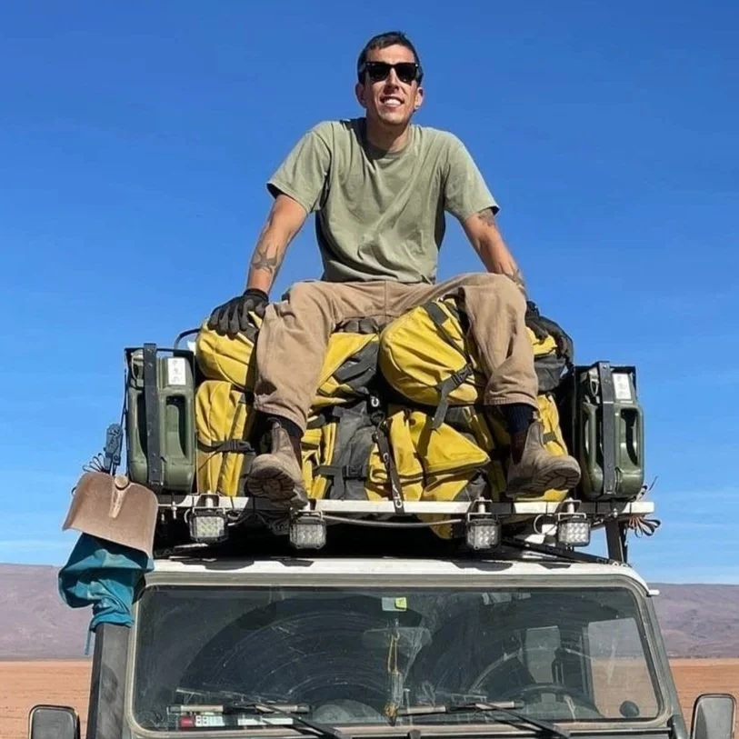 Uomo sorridente seduto sul tetto di un veicolo con molti zaini gialli, in un paesaggio desertico, con cielo blu.
