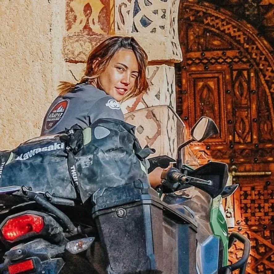 Donna su una moto KTM, con una giacca da moto, davanti a una porta in legno intagliato, che sorride e guarda verso la fotocamera in un ambiente con pareti in pietra decorata.