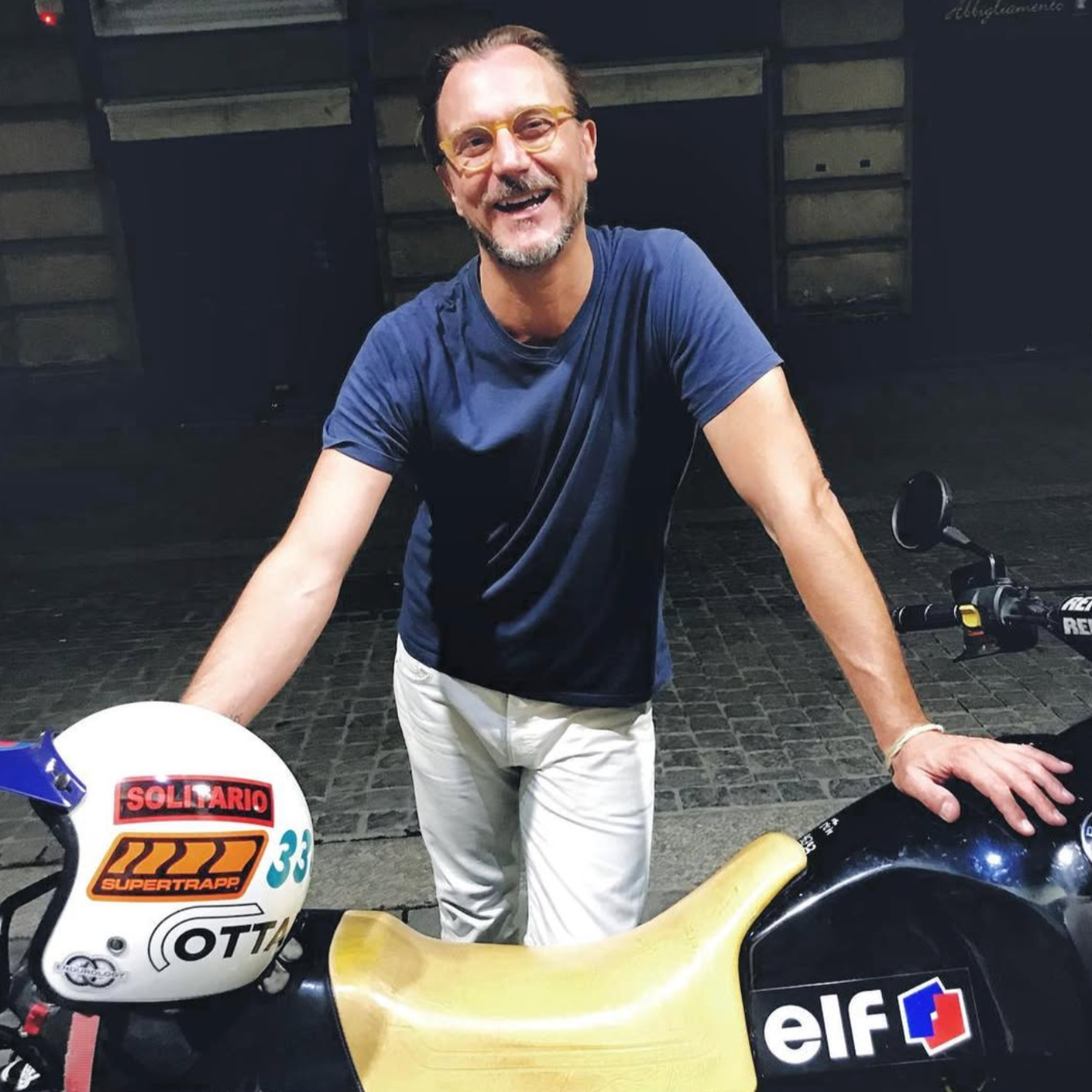 Uomo sorridente con occhiali gialli, maglietta blu, e pantaloni bianchi, appoggiato su una moto da corsa con una casco bianco decorato con vari adesivi.