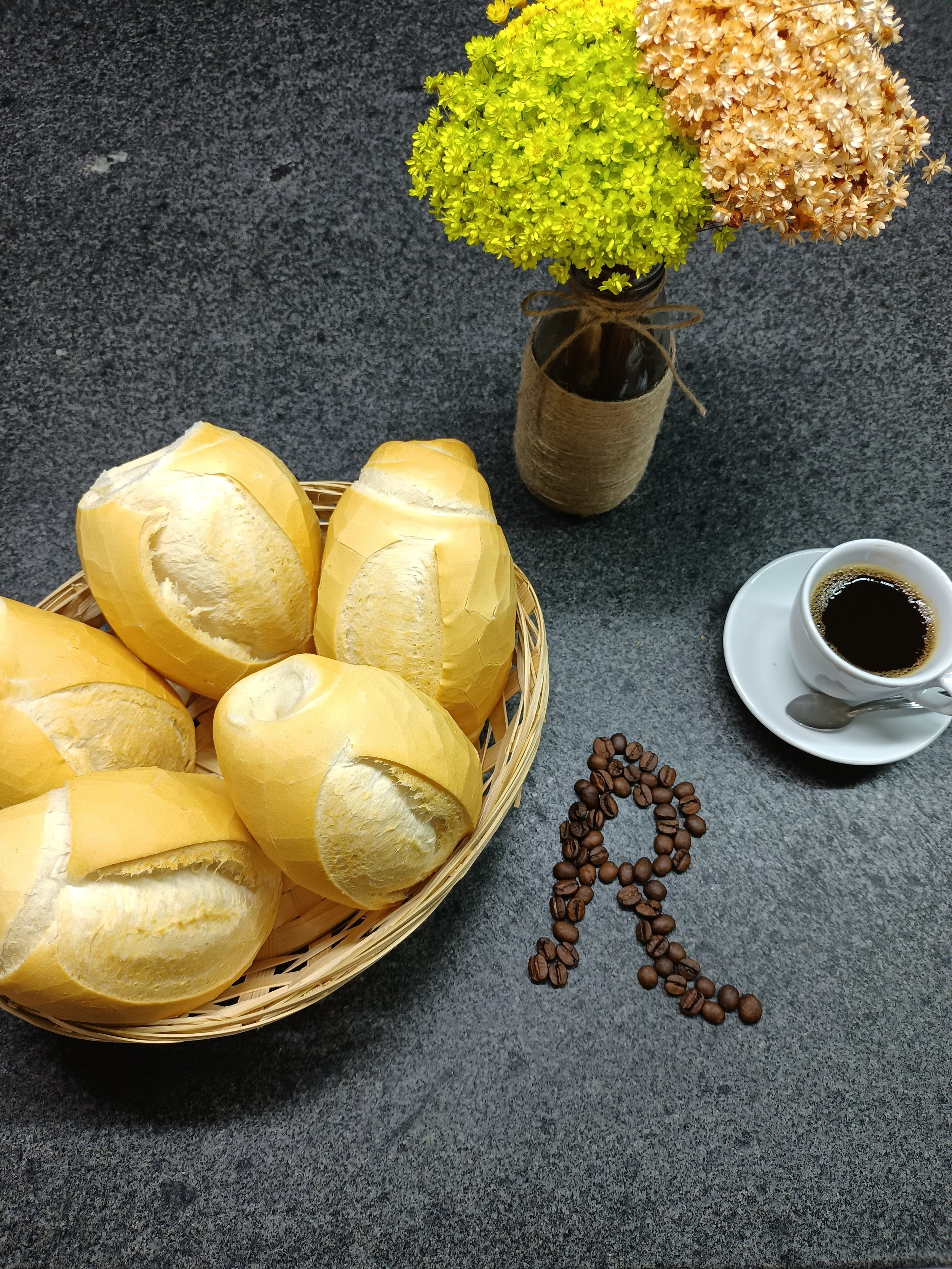 Pães brancos no cesto, copo de café preto, vasos de flores coloridas e sementes de café formando a letra R sobre uma mesa de pedra