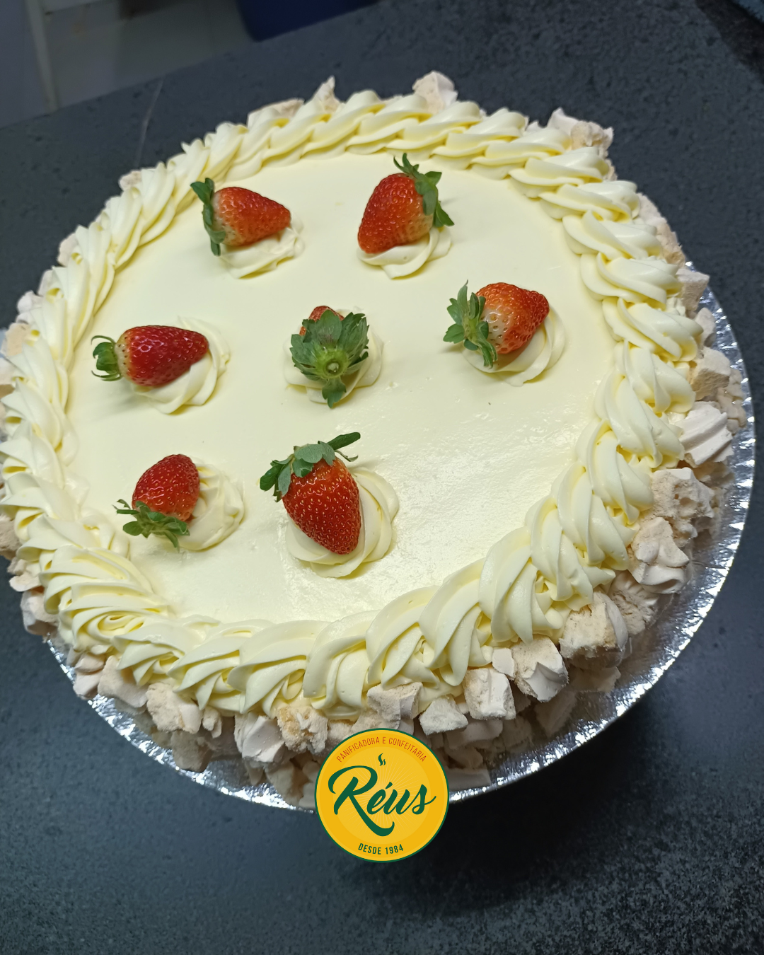 Bolo de aniversário com cobertura de creme branco e morangos frescos decorativos, borda decorada com chantilly e merengues, sobre mesa escura com logotipo de confeitaria Réus na parte inferior