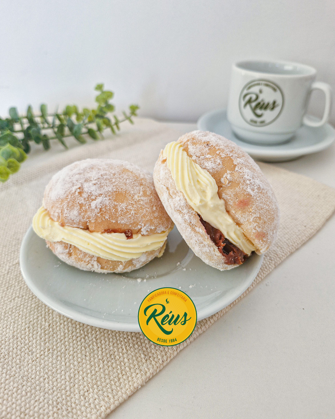 Dois sonhos com recheio de creme de leite e chocolate, cobertos com açúcar e farinha, servidos em prato branco, com xícara de café ao fundo.