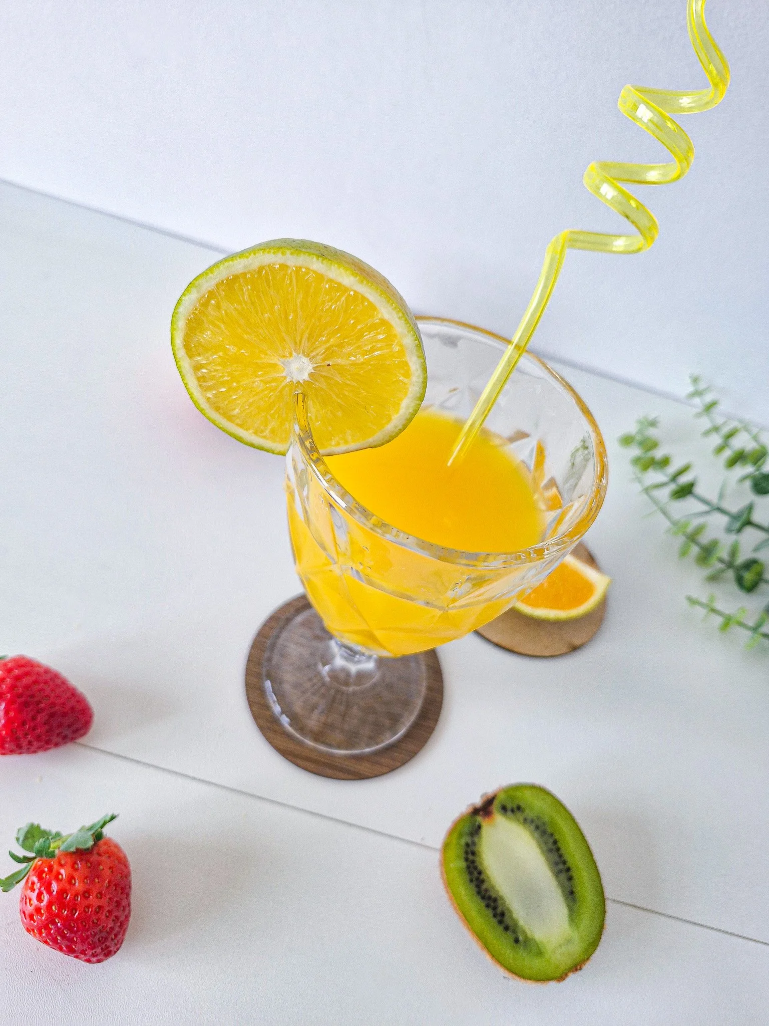 Copo de vidro com uma bebida amarela, decorado com uma fatia de limão na borda e um canudo amarelo ondulado, sobre uma mesa branca com frutas ao redor, incluindo morango, kiwi e uma fatia de laranja.