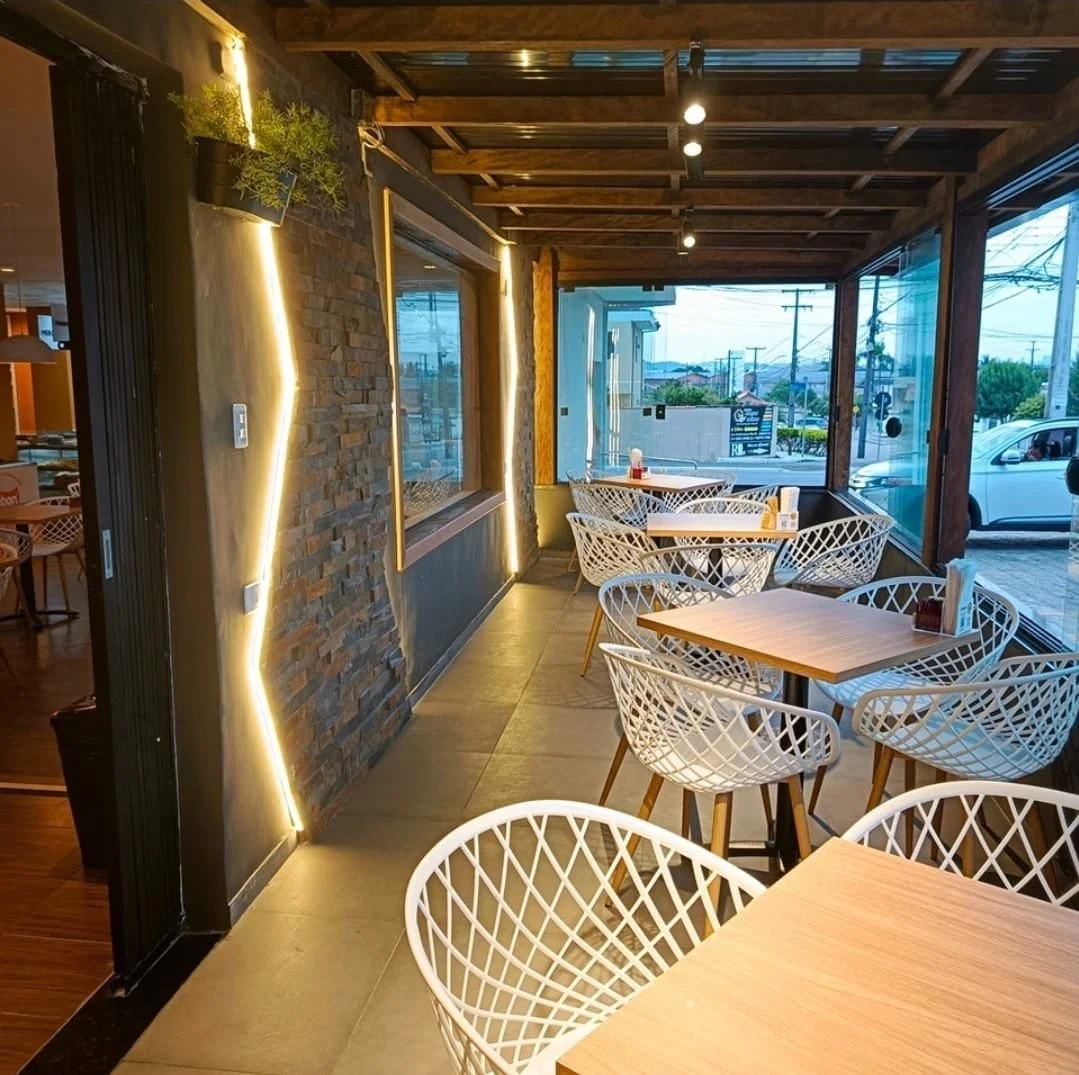 Interior de um restaurante com mesas de madeira e cadeiras brancas de design moderno. Janelas grandes permitem visão do exterior, com carros na rua e postes de energia. Decoração com iluminação suave e parede de pedra com luz de LED.