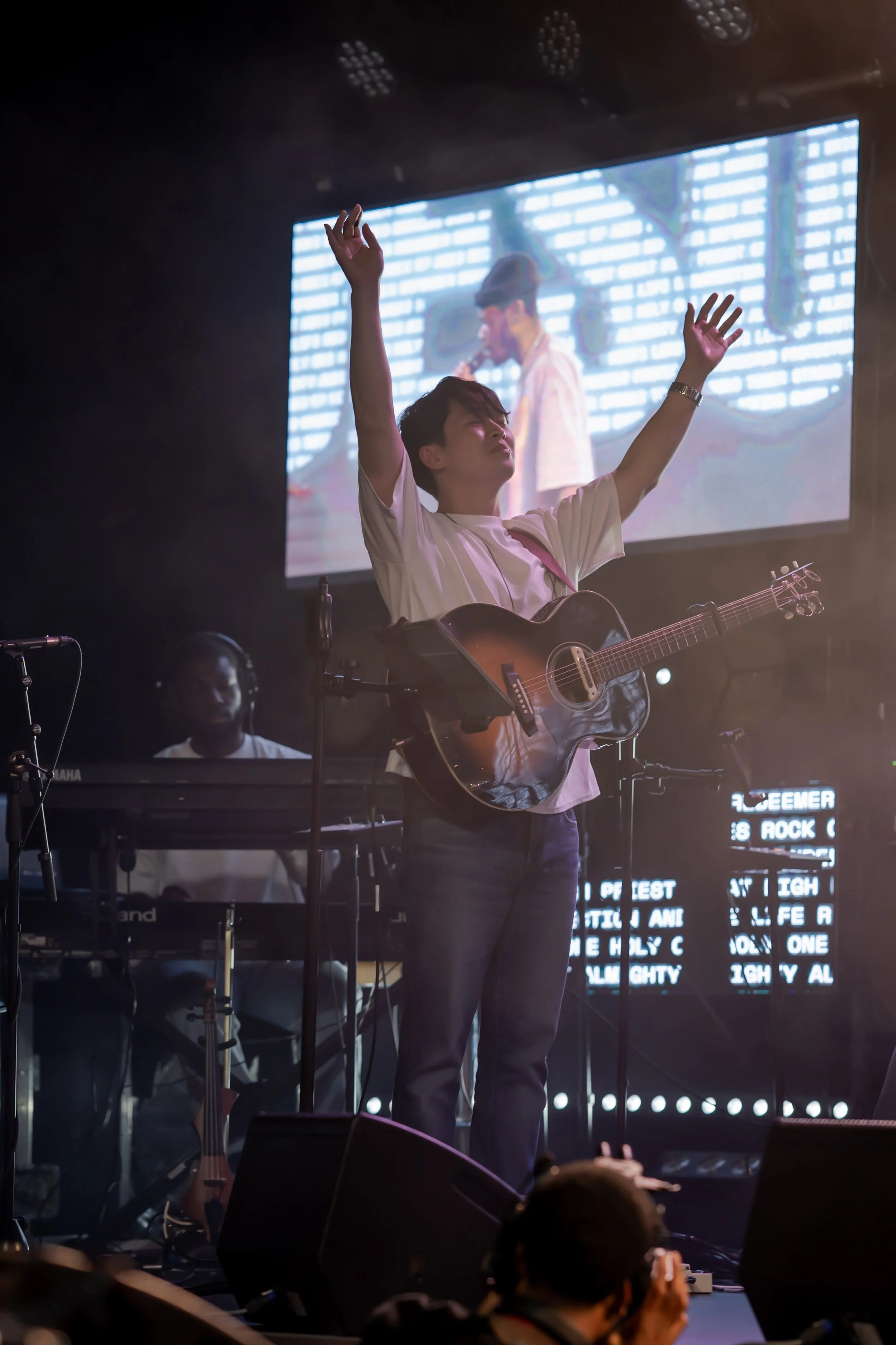 A young man with short dark hair playing an acoustic guitar on stage with eyes closed and arms raised, in front of a group of audience members. A large screen behind him shows a backup performer singing into a microphone. The stage has several musical instruments and equipment, and colorful lights.