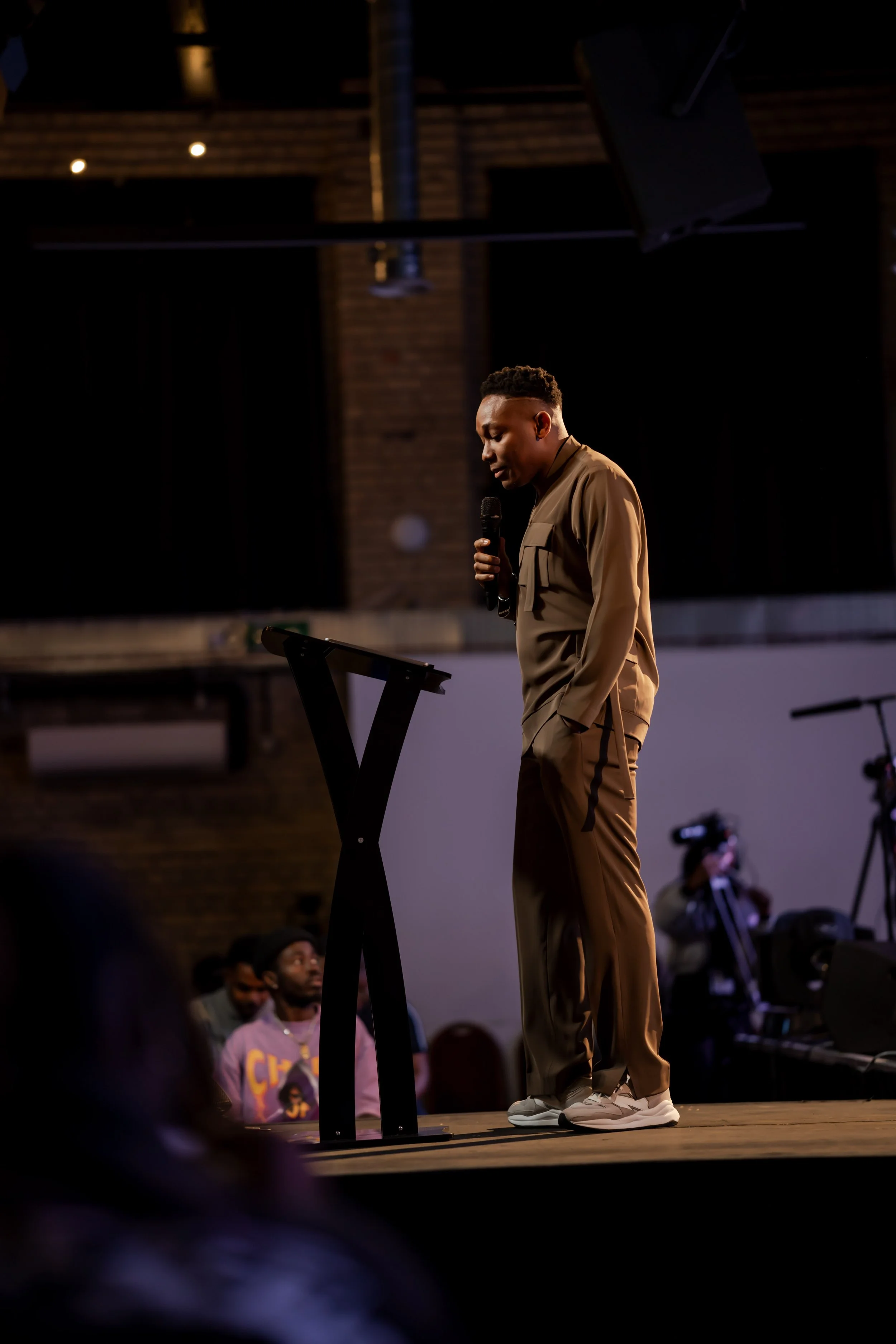 A young man in tan jacket and beige pants stands on stage, holding a microphone and looking down at a music stand in a dimly lit room with a brick wall background. Several people are seated and standing around him, some wearing hoodies and caps, with professional photography equipment visible.