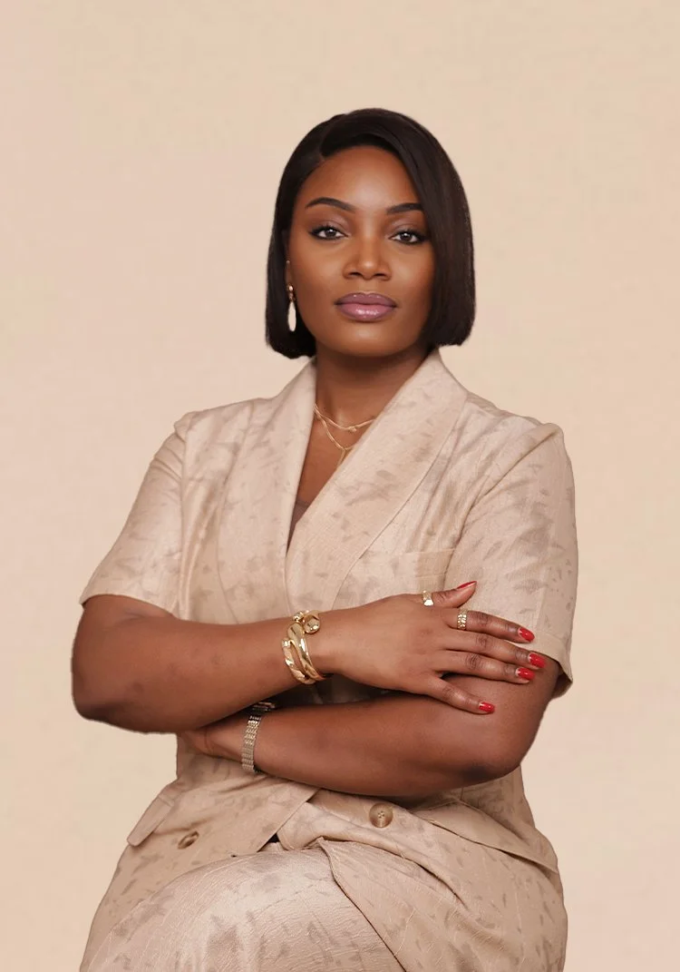 A confident woman with short black hair and medium skin tone wearing a beige suit with jewelry, posing against a plain beige background.