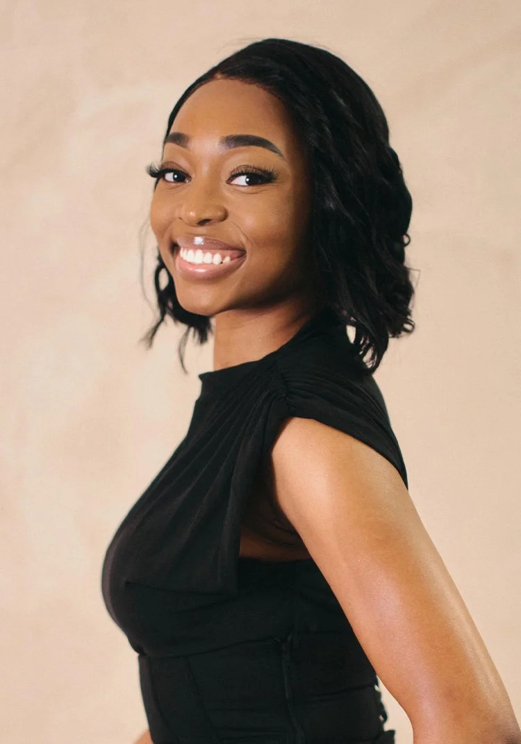 A young woman with shoulder-length curly black hair, smiling, and wearing a black sleeveless top.