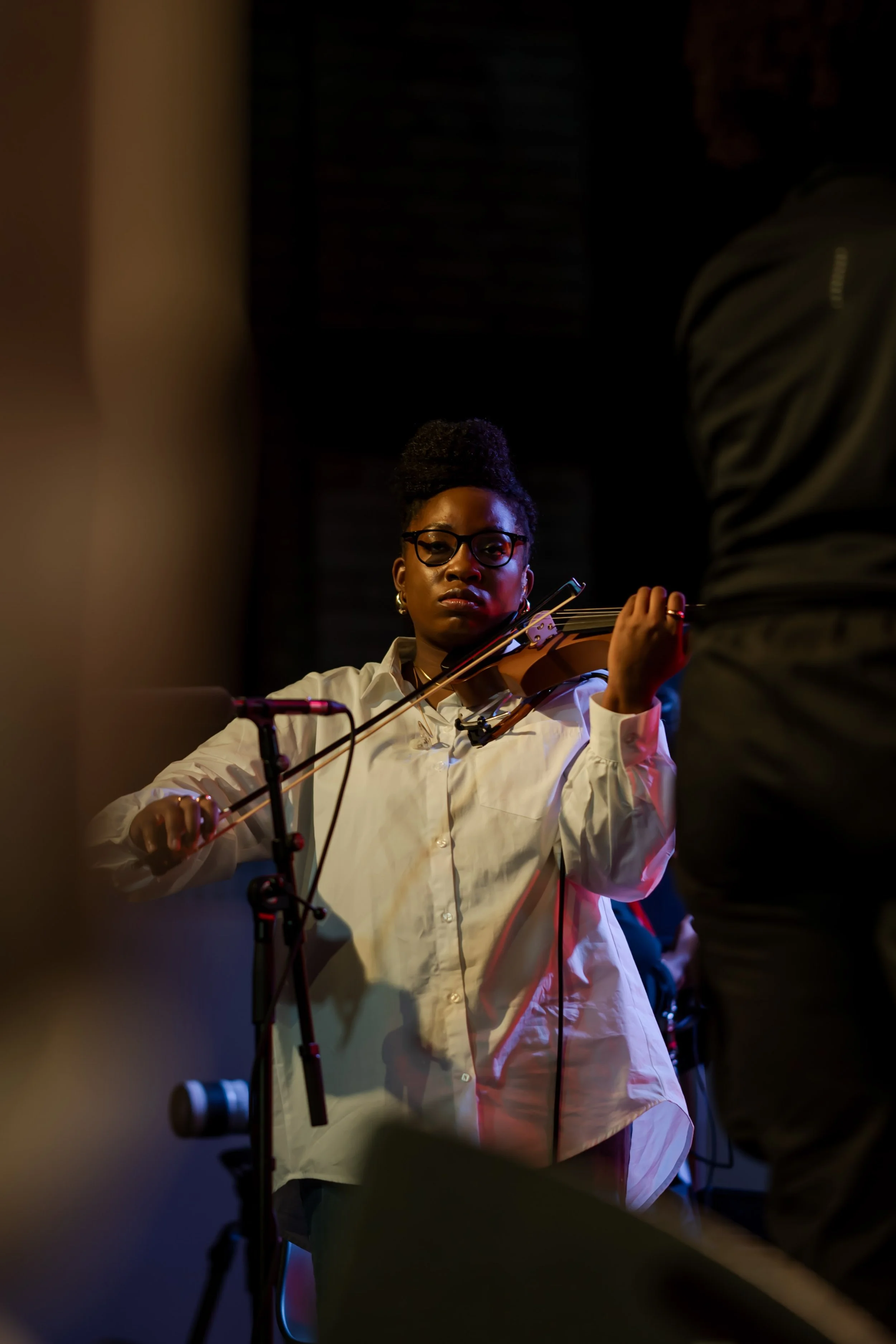 A woman wearing glasses and a white shirt playing the violin in a dark space, with a blurred person in the foreground.