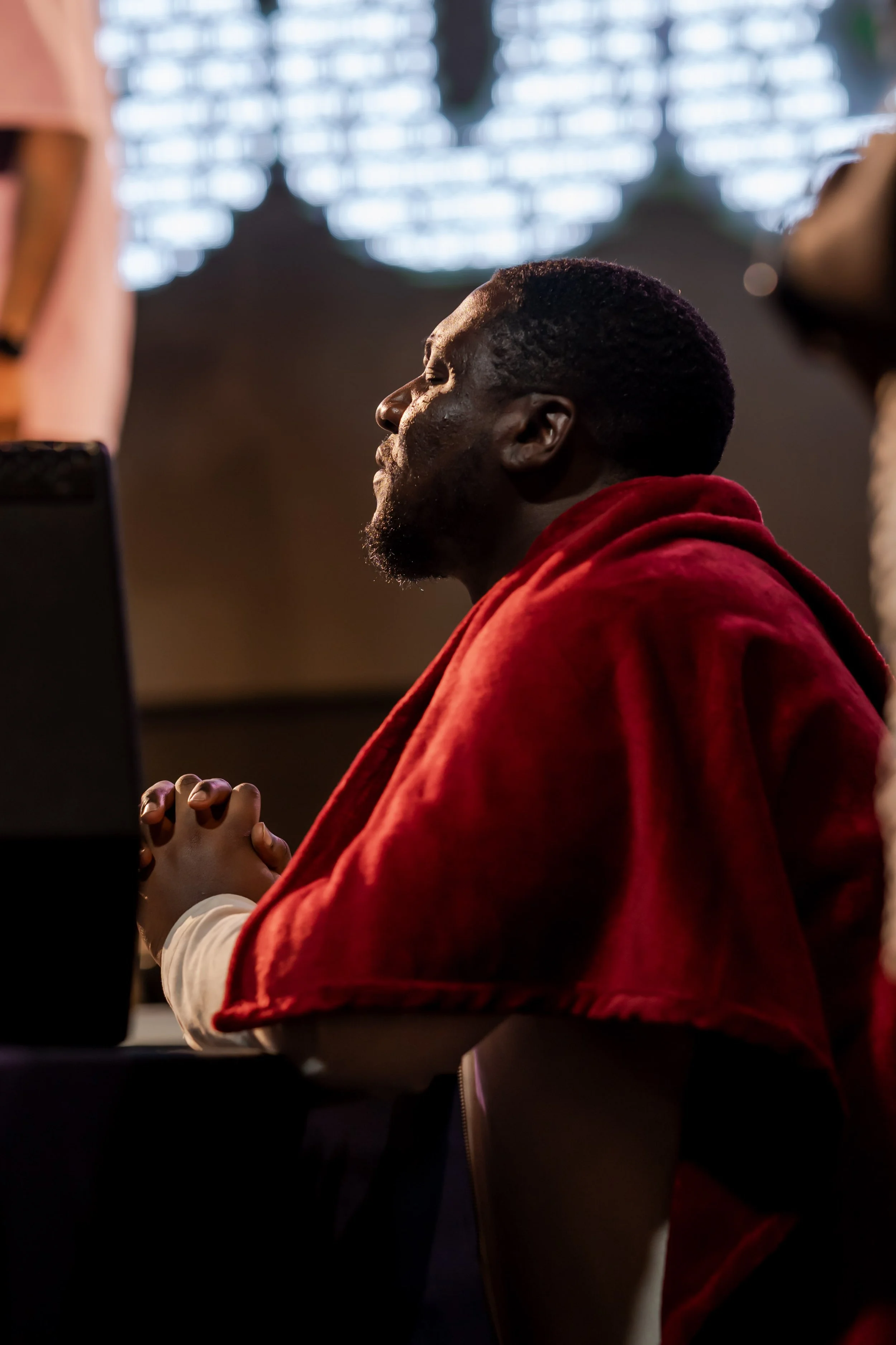 A man in a red hoodie with eyes closed and hands clasped together in prayer or reflection, sitting in a dimly lit indoor setting with sunlight coming through a window in the background.