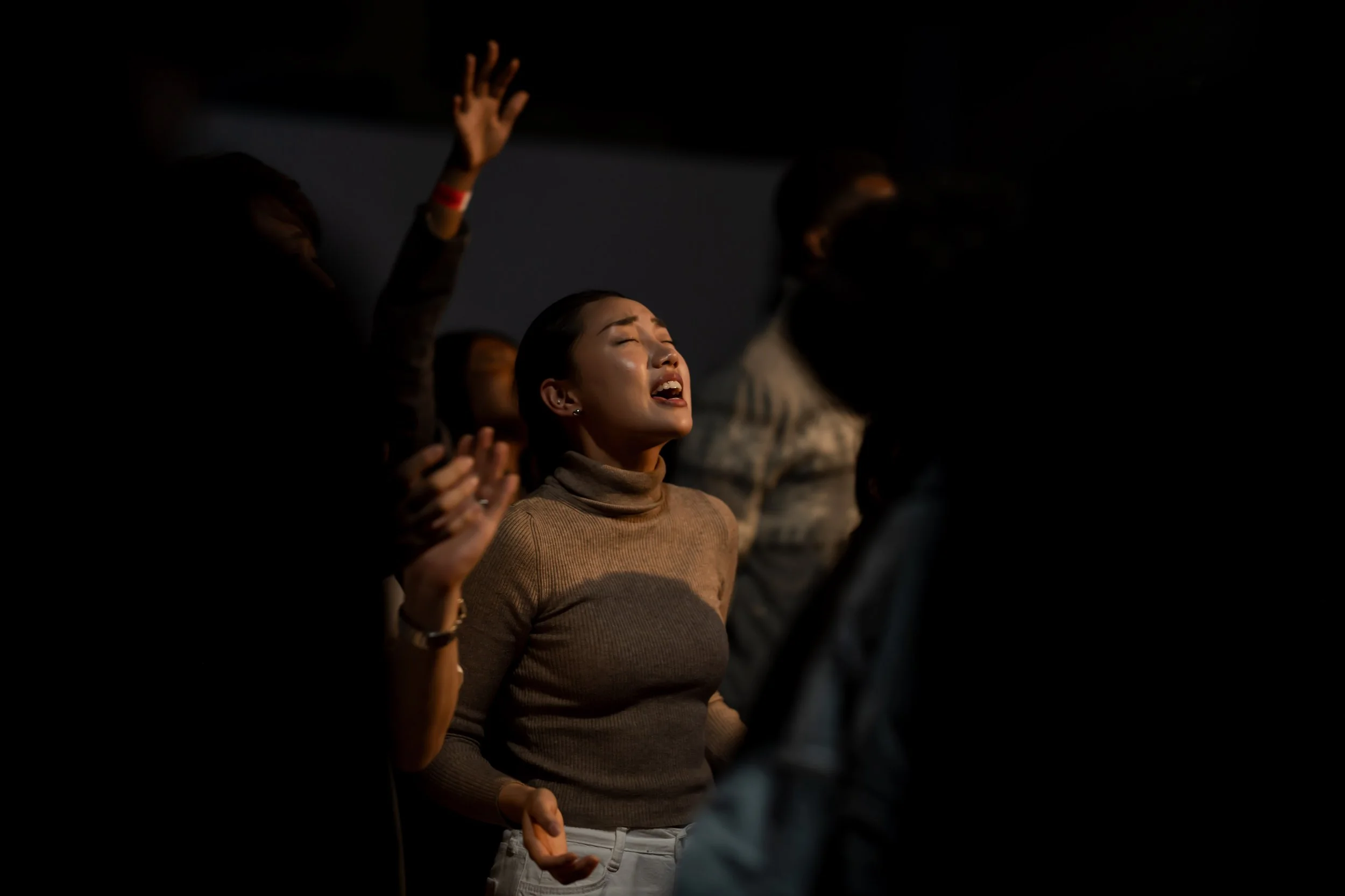 A young woman with dark hair and a beige turtleneck shirt appears to be praying or singing with her eyes closed, surrounded by partially visible individuals in a dark setting.